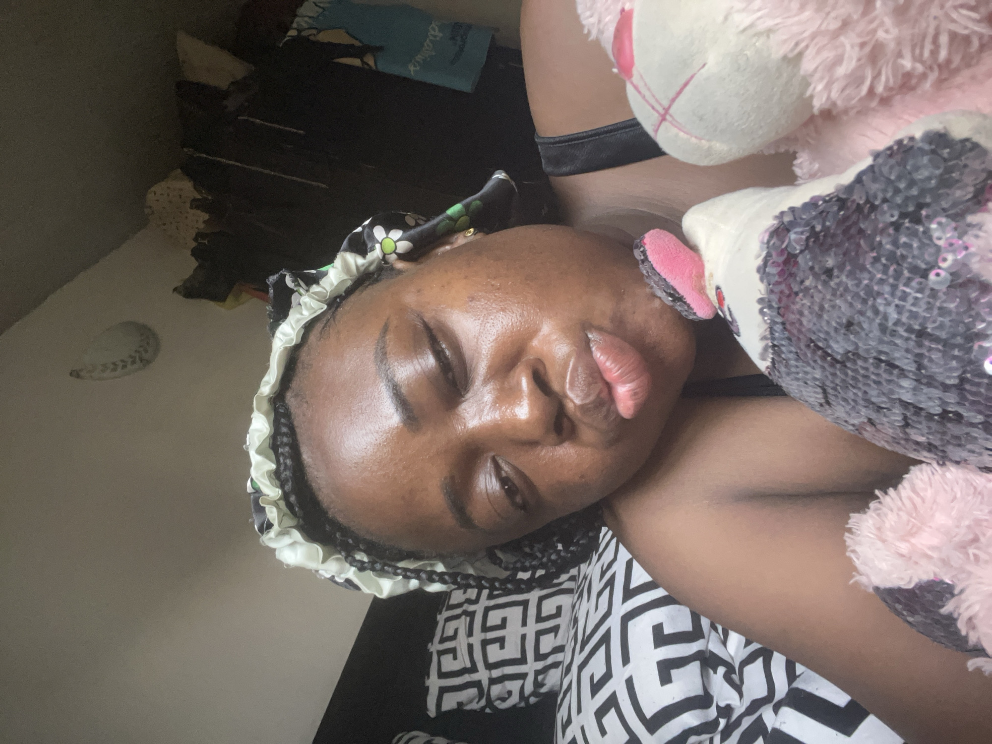 A close-up, angled portrait features a dark-skinned individual, likely a female, lying down indoors, possibly in a bedroom in Abuja, Nigeria. Her eyes are mostly closed, and she displays a playful "duck face" or pouting expression with prominent lips. Her braided hair is partly visible beneath a satin bonnet or hair wrap, which has a black, white, and green floral pattern.
The person's skin shows a natural sheen. She is wearing a dark strap, possibly part of a top or bra. Two plush toys are positioned next to her: one is a fluffy pink toy with a white snout, and the other is adorned with shimmering grey and silver sequins and some pink accents.
The background includes a bed covered with a black and white geometric patterned sheet. A light-colored wall and some dark, indistinct objects, possibly stacked items or furniture, are visible further in the background. The scene suggests a relaxed and intimate moment. There is no visible text in the image.