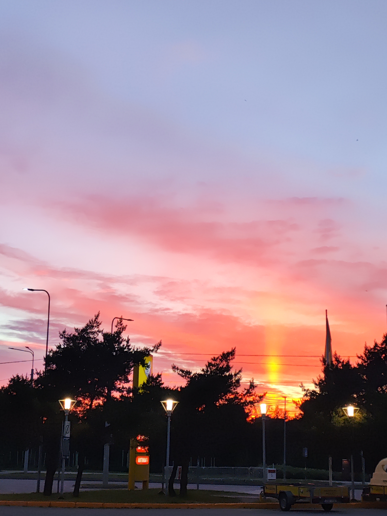 This image captures a vibrant sunset over the city of Tallinn, Estonia. The sky is ablaze with hues of pink, orange, and yellow, with streaks of purple and blue higher up, creating a dramatic backdrop. Silhouetted against this colorful sky are the dark shapes of trees, some with their branches reaching upwards. Several streetlights are visible, casting a warm glow. In the foreground, a trailer with yellow accents sits on a paved surface. A yellow sign with red lettering, likely indicating an automated service, is also present. The scene appears to be an outdoor public space, possibly a parking lot or roadside area, given the streetlights, pavement, and trailer. The overall impression is one of natural beauty juxtaposed with urban elements at the close of day.