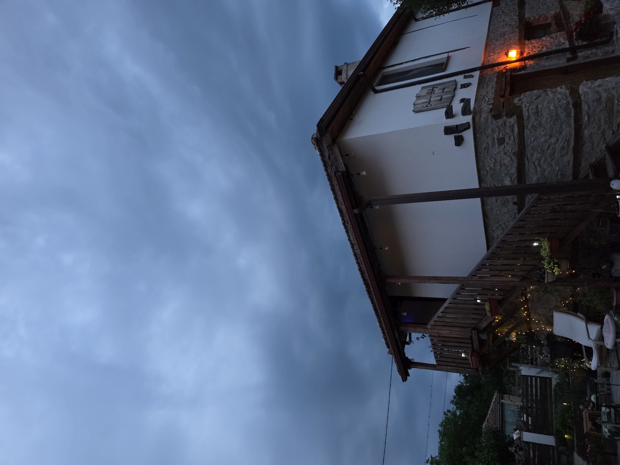 A slightly angled, full-shot image depicts a white house with a stone foundation on the right side of the frame, against a dramatic, cloudy, dusky sky. The house has a wooden porch with a staircase leading up to it, decorated with string lights and potted plants. There is a small orange lantern illuminating a doorway to the right of the frame. The house has a red-tiled roof, and a wooden sign that reads "Emanouil Idzof Guest House" is mounted on the wall. To the left of the house, there are more buildings with red roofs and trees. The overall scene suggests a rural or village setting at twilight, possibly indicating a coming storm or the end of the day. There are no people visible in the image.