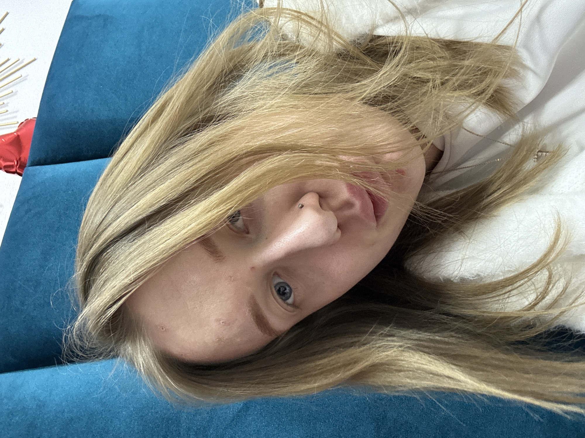 A woman with blonde hair and blue eyes lies on a blue sofa with a white object nearby. She has a nose piercing. There is no information to suggest this image was taken in Rostov-on-Don, Russia.