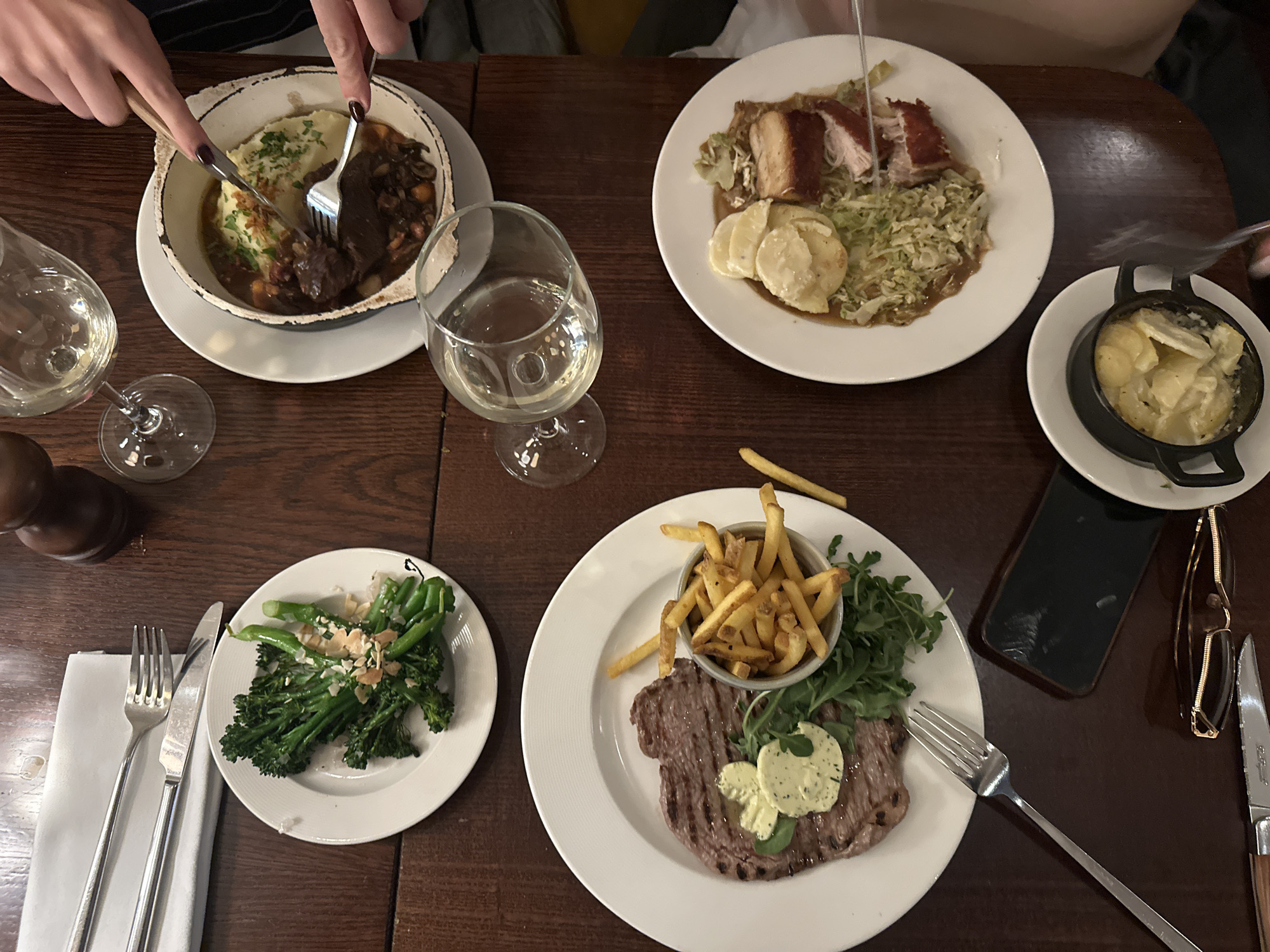 The image shows a table at a restaurant, likely in St Albans, UK, based on the style and types of dishes. There are at least four people dining, evident from the various plates of food and hands visible. Dishes include:
* A stew or casserole with mashed potatoes in a distressed white bowl.
* Pork belly with shredded cabbage and potatoes.
* Broccolini topped with nuts.
* A steak with fries served in a metal cup and arugula.
* A dish of sliced potatoes, possibly dauphinoise.
Each place setting has silverware. There are also glasses of white wine on the table. A smartphone and glasses case are placed on the table next to one of the plates. The table itself is a dark, polished wood. The people are in the process of eating, as forks are in motion and some food has already been consumed.
