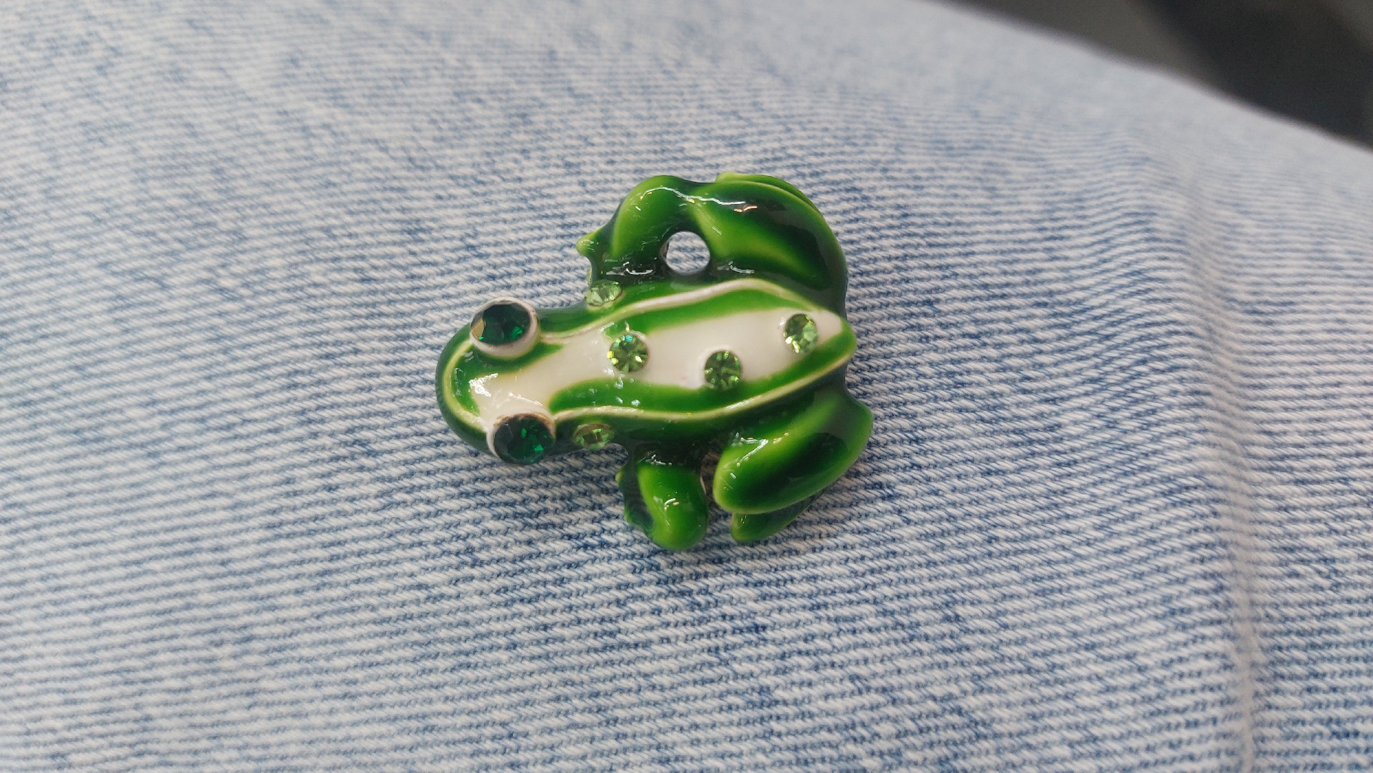 A green and white frog-shaped brooch with green rhinestones sits on a textured light blue fabric.