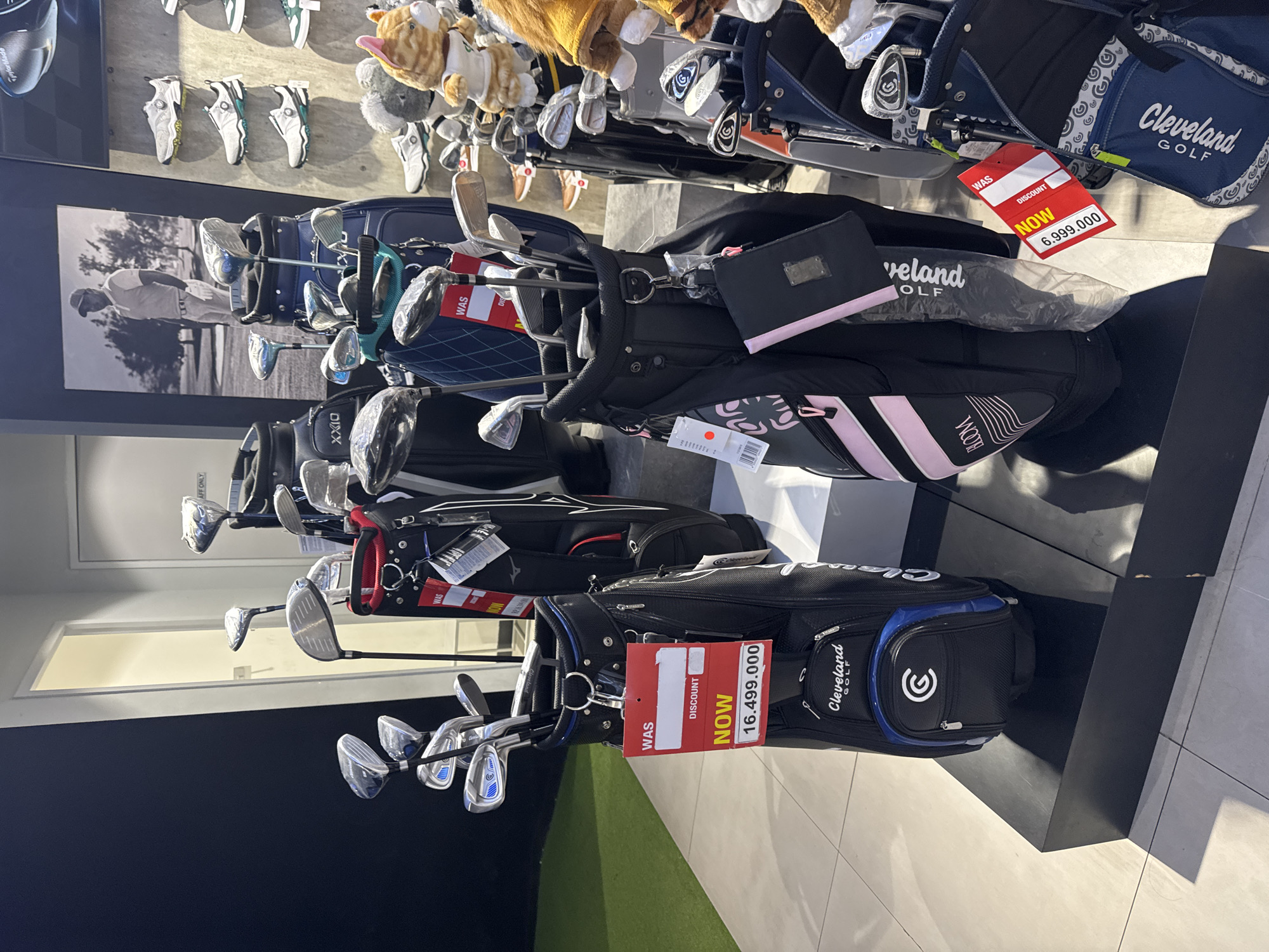 The image depicts the interior of a golf pro shop, likely located in Kuta, Indonesia, as suggested by a sign with "Kuta" and "Indonesia" in the image. The scene is filled with various golf equipment, primarily golf bags and clubs, displayed on tiered platforms and shelves.
In the foreground, two golf bags are prominently featured. The bag on the left is black with blue accents and the "Cleveland Golf" logo. It contains a set of golf clubs. A red discount tag is attached to this bag, displaying "WAS" and "DISCOUNT" with a "NOW" price of "16.499.000". The bag on the right is a black and pink "XXIO" branded bag, also holding golf clubs. It has a smaller red discount tag indicating a "NOW" price of "6.999.000".
Behind these bags, more golf bags and clubs are visible, along with shelves displaying golf shoes and stuffed animal mascots. A large black and white photograph of a golfer is mounted on the wall behind the display. The environment appears well-lit with artificial lighting. There are no people visible in the image. The floor is tiled.