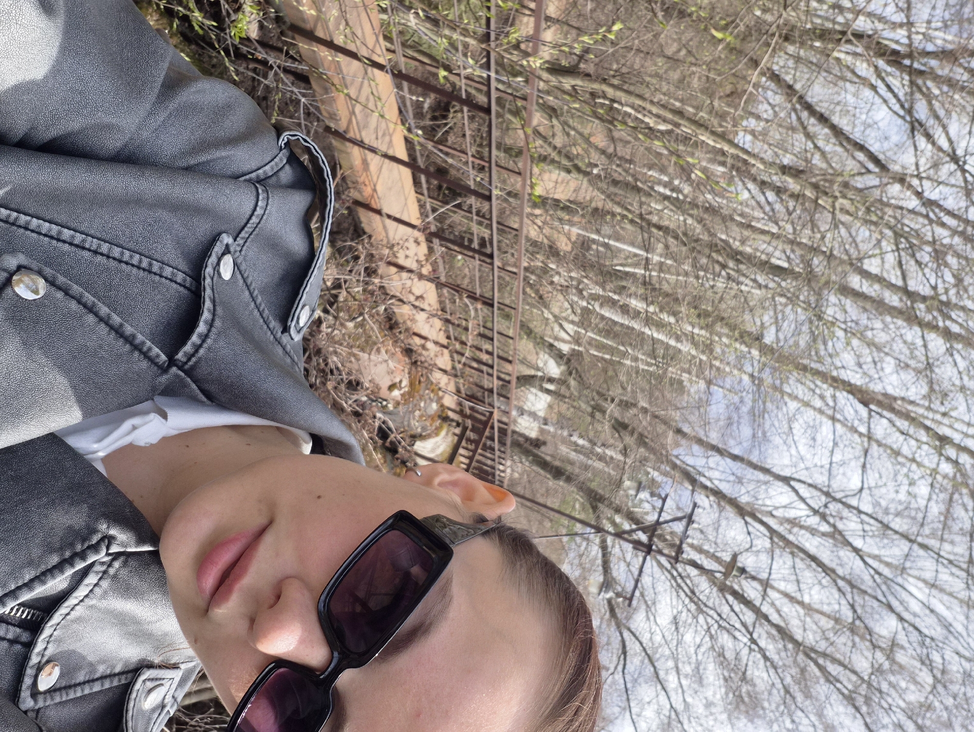 An outdoor portrait shot shows a young person in the foreground, angled to the left, capturing their head, neck, and upper chest. They are wearing a dark grey or black leather-like jacket with metallic buttons over a white top. Dark rectangular sunglasses obscure their eyes, while their lips are slightly parted. Their short, light brown hair is visible.
The background is an out-of-focus natural setting, suggesting late winter or early spring in Chaikovsky, Russia. Numerous bare tree branches, some with nascent light green buds, stretch upwards towards a bright, hazy sky. A rusty, dark metal structure, possibly a fence or part of a bridge, runs horizontally across the mid-ground behind the subject. The ground visible near the structure is dry and earthy. The overall lighting indicates it is daytime. The person appears calm and relaxed. No text is visible in the image.