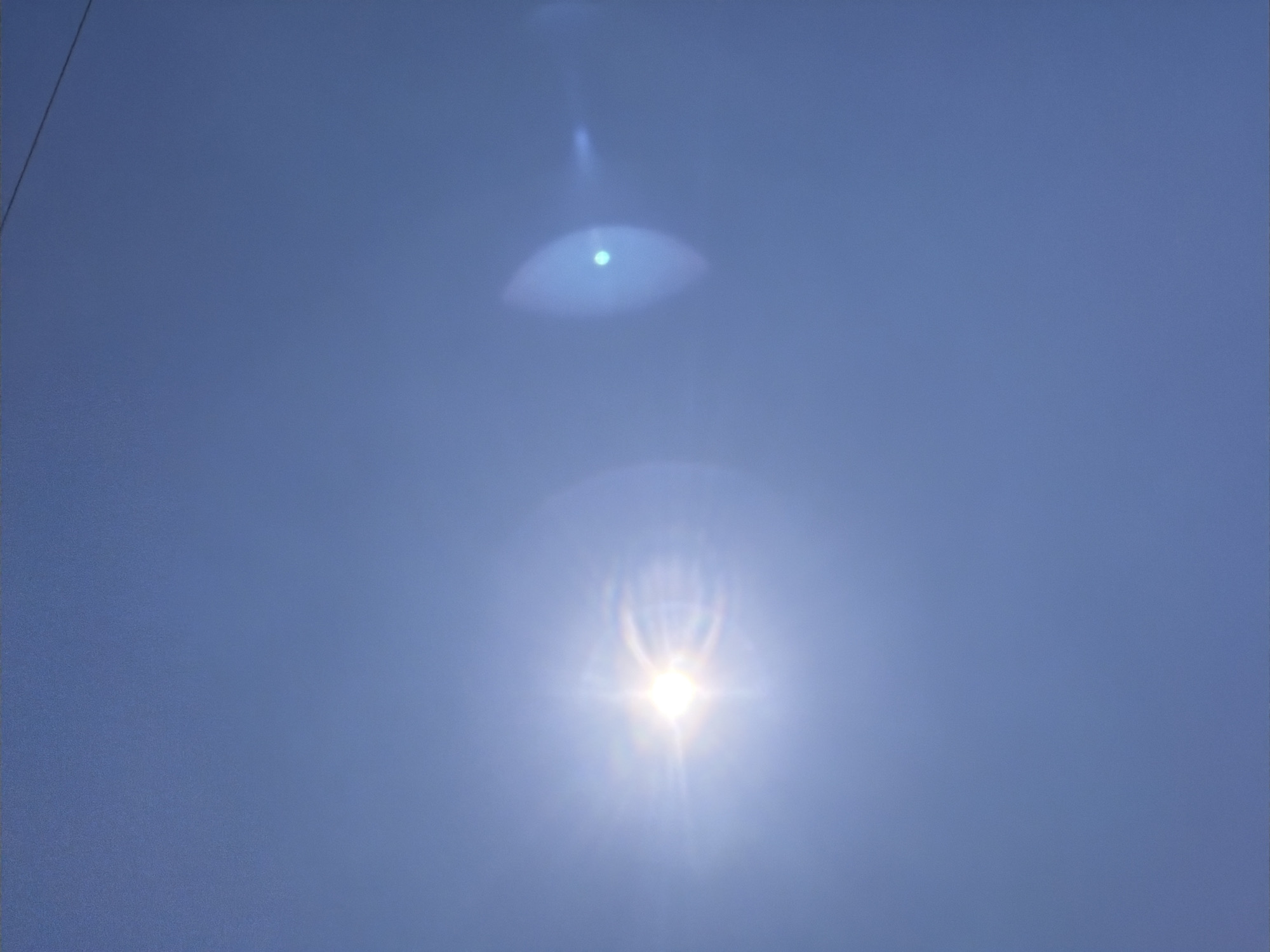 A clear, blue sky fills the frame, with the bright sun positioned in the upper left quadrant. Lens flare emanating from the sun creates a starburst effect and a fainter, circular artifact to the right. A thin, dark wire is visible against the sky in the upper right corner, suggesting it is suspended overhead. The scene is devoid of any recognizable landmarks or structures of Ust'-Dzhigutinka, Russia.