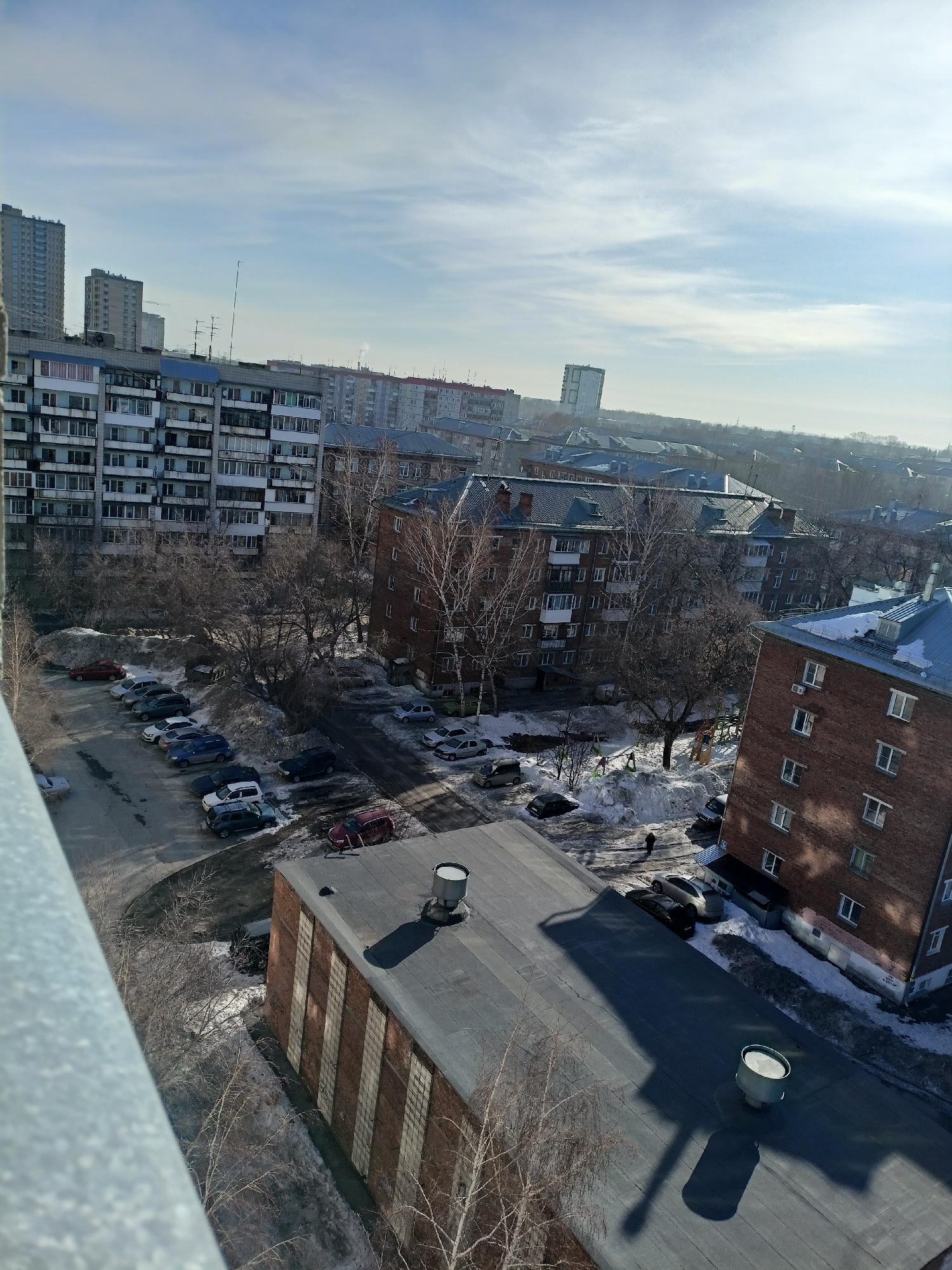 The photograph captures an elevated view of a residential neighborhood in Novosibirsk, Russia, during a wintery day with a partially cloudy sky. The foreground includes a section of a building exterior, possibly a balcony, partially obscured by bare branches of a tree.
A parking area, visible on the left, is filled with a variety of parked cars, some covered in snow. Mid-rise apartment buildings, constructed with gray concrete and brick exteriors, dominate the mid-ground. Patches of snow are evident on rooftops and on the ground in shaded areas, indicating recent snowfall. A small playground area near one of the buildings appears to be covered in snow. Distant buildings and light-colored sky create a hazy horizon. Two people walk near the playground.
The lighting suggests the image was taken during the morning or afternoon. The scene is typical for a Russian urban landscape in winter.
