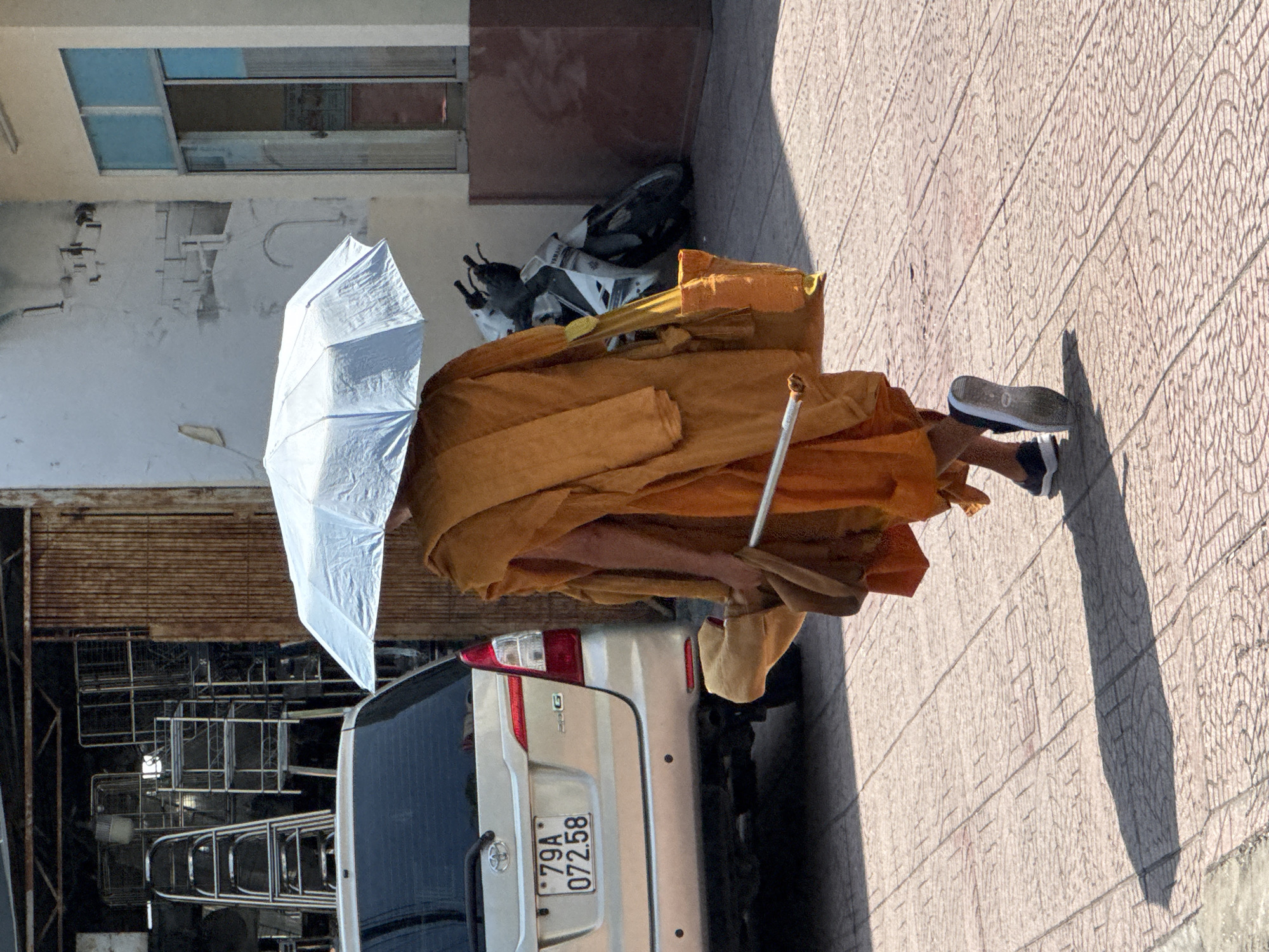A Buddhist monk walks away from the camera, holding a light blue umbrella over their head. The monk is wearing an orange robe and carrying a tan bag over their right shoulder. The monk is also holding a silver pole in their right hand. In the background, there is a silver Toyota Innova car with a license plate reading "79A 072.58". Next to the car is a white and black Yamaha motorcycle. The scene appears to be on a paved walkway in front of a building with a large metal door. The overall environment suggests an urban setting in Nha Trang, Vietnam, possibly a market or a street in a commercial area, given the presence of the car, motorcycle, and what appears to be storage units or shelves in the background.