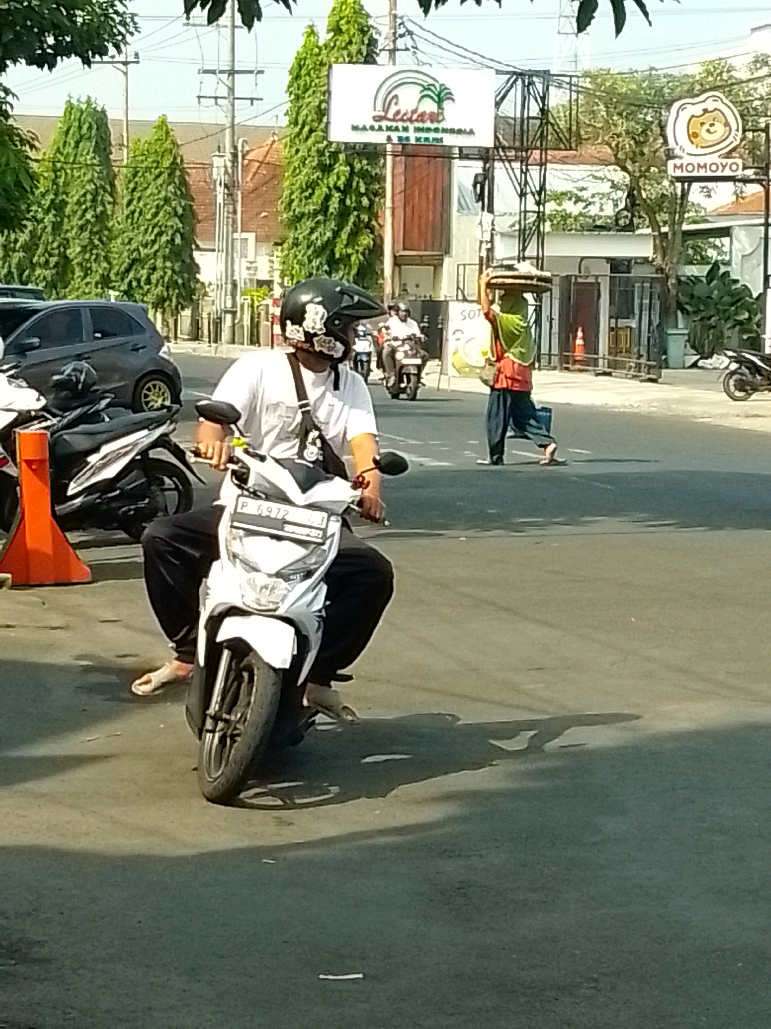 The image captures a street scene in Jember, Indonesia, likely in the daytime given the bright lighting and shadows. In the foreground, a person is riding a white scooter, wearing a white t-shirt, black pants, and a black helmet adorned with white floral stickers. They have a dark bag slung across their chest. The scooter has the license plate "P 6972 GJ".
In the middle ground, a person is walking across the street, carrying a circular tray laden with goods on their head, covered by a green cloth. They are wearing an orange top and blue pants. Behind this person, a billboard with text and an image of a cartoon lion is visible, identified as "MOMOYO". Another billboard with palm trees and text is also present.
Further back, several other motorbikes and a dark gray car are visible. The environment appears to be a street with paved roads, sidewalks, and some vegetation, including tall, slender trees. Utility poles and wires are also present, indicating an urban setting. The overall atmosphere suggests a busy street in a commercial or residential area of Jember.