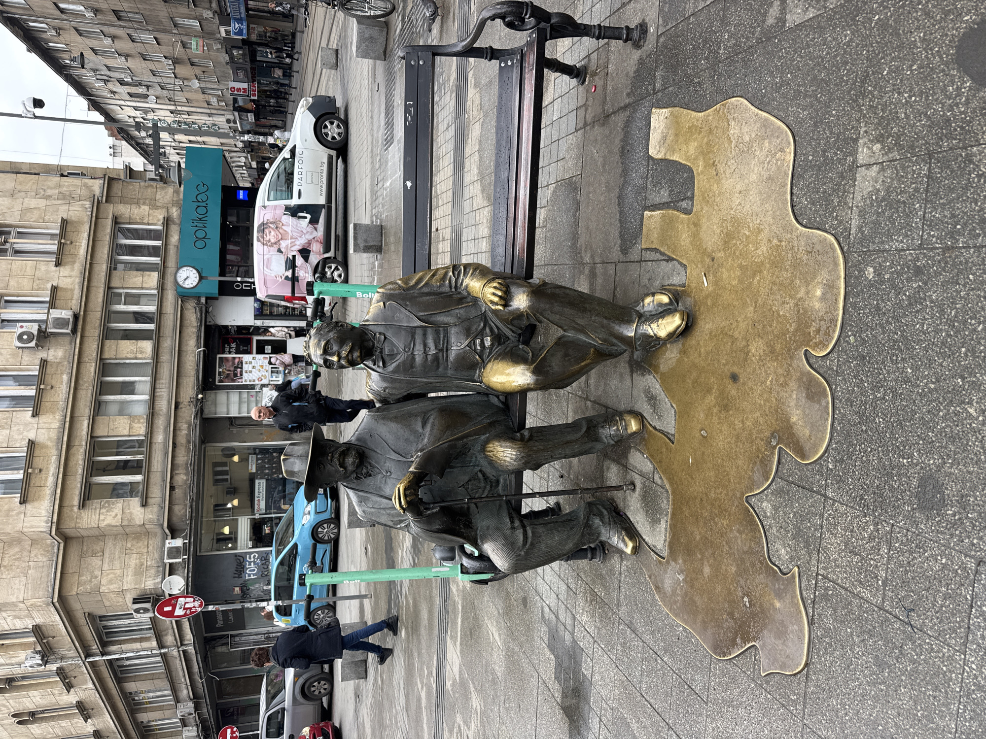 The image depicts a bronze sculpture of two seated men on a bench in Sofia, Bulgaria. The sculpture is located on a paved sidewalk in front of a building with various shops and businesses.
The two men are dressed in formal attire and appear to be deep in conversation. One of the men is wearing a hat and holding a cane. A bronze outline of Bulgaria surrounds the sculpture.
Several people are walking by in the background, including a man in a black jacket and an older gentleman. Various cars and e-scooters are visible in the scene, including a white van advertising "optikabc". Street signs are posted nearby. The sky is overcast.