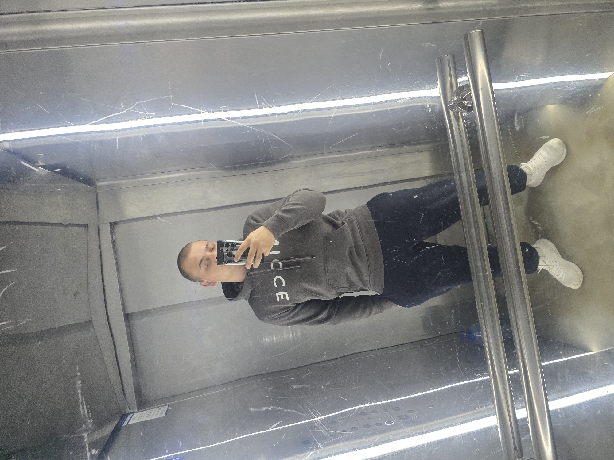 The image depicts a reflection of a person taking a selfie in an elevator. The person is wearing a gray hoodie with the word "NICE" printed on it, dark pants, and white sneakers. The elevator has metal walls with scratches and a metal handrail. The location is implied to be in the city of Sofia, Bulgaria.