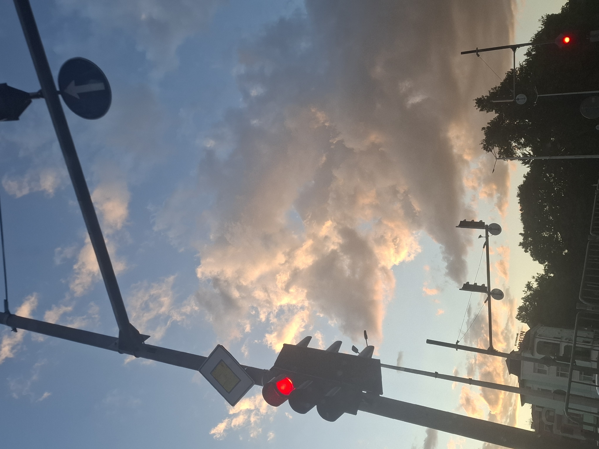 A close-up, low-angle shot captures a traffic light with a red light illuminated, silhouetted against a sky filled with dramatic clouds. The clouds are illuminated by the setting or rising sun, casting warm orange and pink hues across their surfaces, contrasting with the cooler blue tones of the sky. The traffic light is mounted on a metal pole, accompanied by a diamond-shaped road sign with a yellow interior. Further in the background, a horizontal street light bar extends across the frame, supporting additional traffic lights and circular signs with white arrows pointing upwards. The scene is partially obscured by trees and buildings, suggesting an urban environment. The overall impression is one of an intersection at dusk or dawn, with the red light indicating a stop.