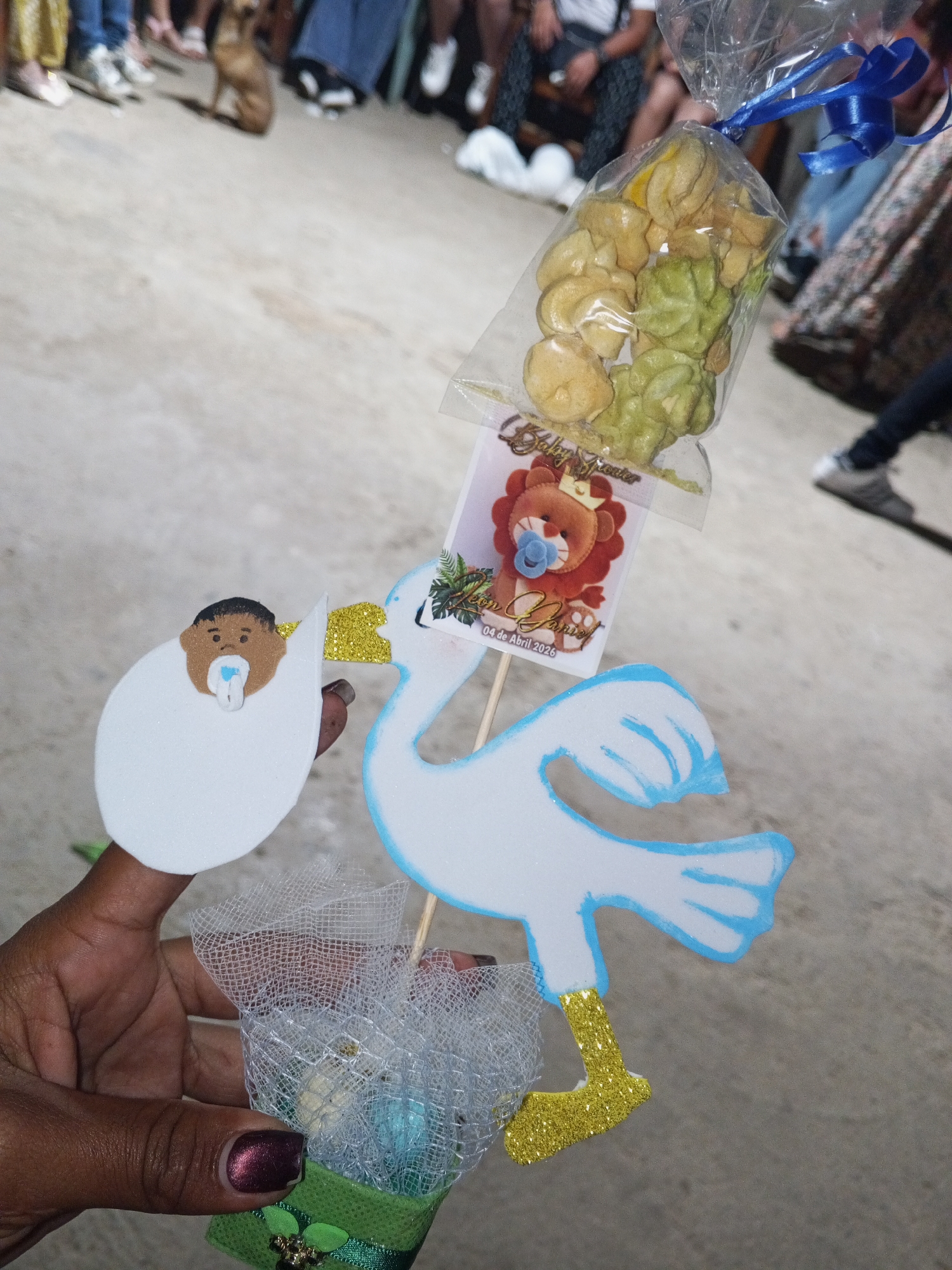 Here is a detailed description of the image:
A hand holds a baby shower favor that is a stork delivering a baby. The favor is composed of several elements: A white foam stork cutout with blue edging and gold glitter feet, carrying a foam baby cutout. The baby has brown skin and a blue pacifier. There is also a card attached featuring a lion design and the text "Baby Shower" and "Leon Daniel, 04 de Abril 2026." Above the card, there is a small clear plastic bag tied with a blue ribbon, containing light yellow and green candies. The stork and baby are supported on a wooden skewer stick, and the base of the favor is wrapped in a green decorative ribbon and netting, possibly containing small items or treats.
The hand holding the favor has dark skin and dark nail polish. The background shows a gathering of people; some are seated, and some are standing. A dog can be seen behind the crowd. The location is not discernible from the photograph.