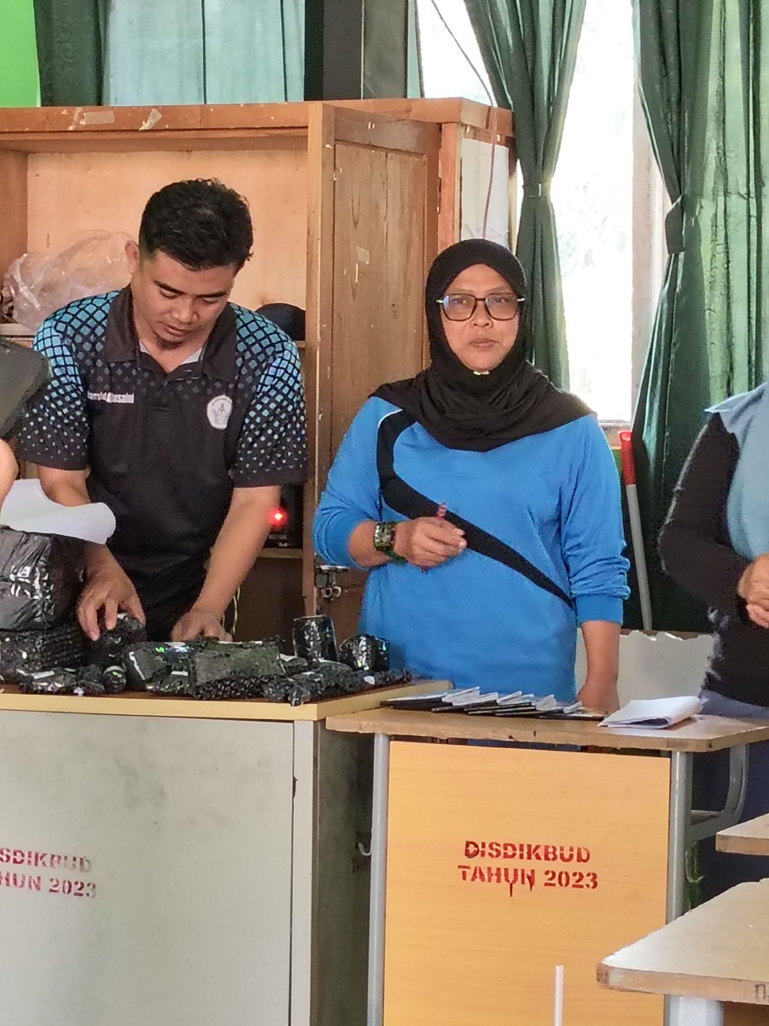 In the city of Lori, Indonesia, two individuals are present. A man wearing a black polo shirt with a blue and white checkered pattern is standing behind a counter, looking down at packages he is handling. To his right, a woman wearing a blue long-sleeved shirt and a black headscarf is also standing behind the counter, looking forward. She is wearing glasses and holding a red pen. Several packages wrapped in black bubble wrap are spread across the counter. Behind the counter, there are shelves filled with various items, and green curtains hang in the background. One of the counters has "DISDIKBUD TAHUN 2023" stenciled in red letters.