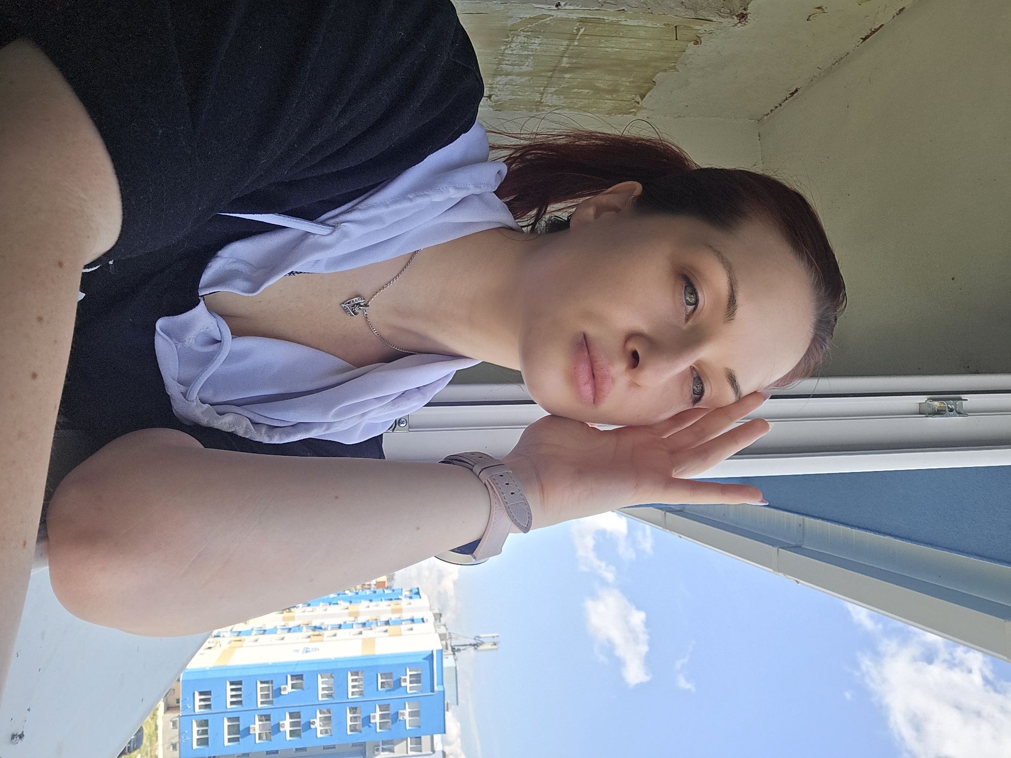 A young woman is captured in a close-up, selfie-style shot, taken from what appears to be a balcony or window ledge in Burgas, Bulgaria. She is positioned on her side, looking directly at the camera with a calm and slightly pensive expression. Her left hand is gently resting near her cheek. She is wearing a black shirt with a light-colored, possibly pale blue or white, hooded top underneath, and a silver chain necklace featuring a heart-shaped pendant. A light-colored watch is visible on her left wrist. Her hair, which appears dark, is pulled back from her face.
The background reveals a bright, clear blue sky with a few scattered white clouds, indicative of sunny weather during daylight hours. Below the sky, an urban landscape is visible, featuring a prominent multi-story building painted in blue and white, characteristic of residential architecture. The wall or ceiling above her head, closer to the camera, is light-colored and shows texture, suggesting an unfinished or aged plaster surface. There is no visible text in the image.