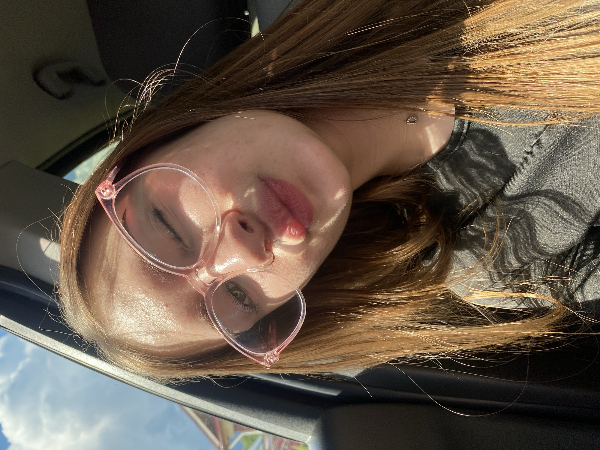A young Caucasian woman with long, light brown hair and fair skin is seen in a close-up selfie taken from a slightly low angle, presumably inside a vehicle. She is wearing pink-rimmed glasses and has her mouth puckered as if blowing a kiss, with her tongue slightly visible. Sunlight is hitting her face, creating highlights and shadows, and her left eye is slightly squinted. She is wearing a black shirt, and a delicate necklace with a small, sparkling 'D' pendant is visible on her collarbone. The background suggests an outdoor environment with a blue sky and clouds visible through a window, and the interior of a car, including a dark gray seat and a portion of the car door frame. The woman's hair is slightly windblown, with strands catching the sunlight. There is no indication of her location within the city of Dyakovo, Russia.