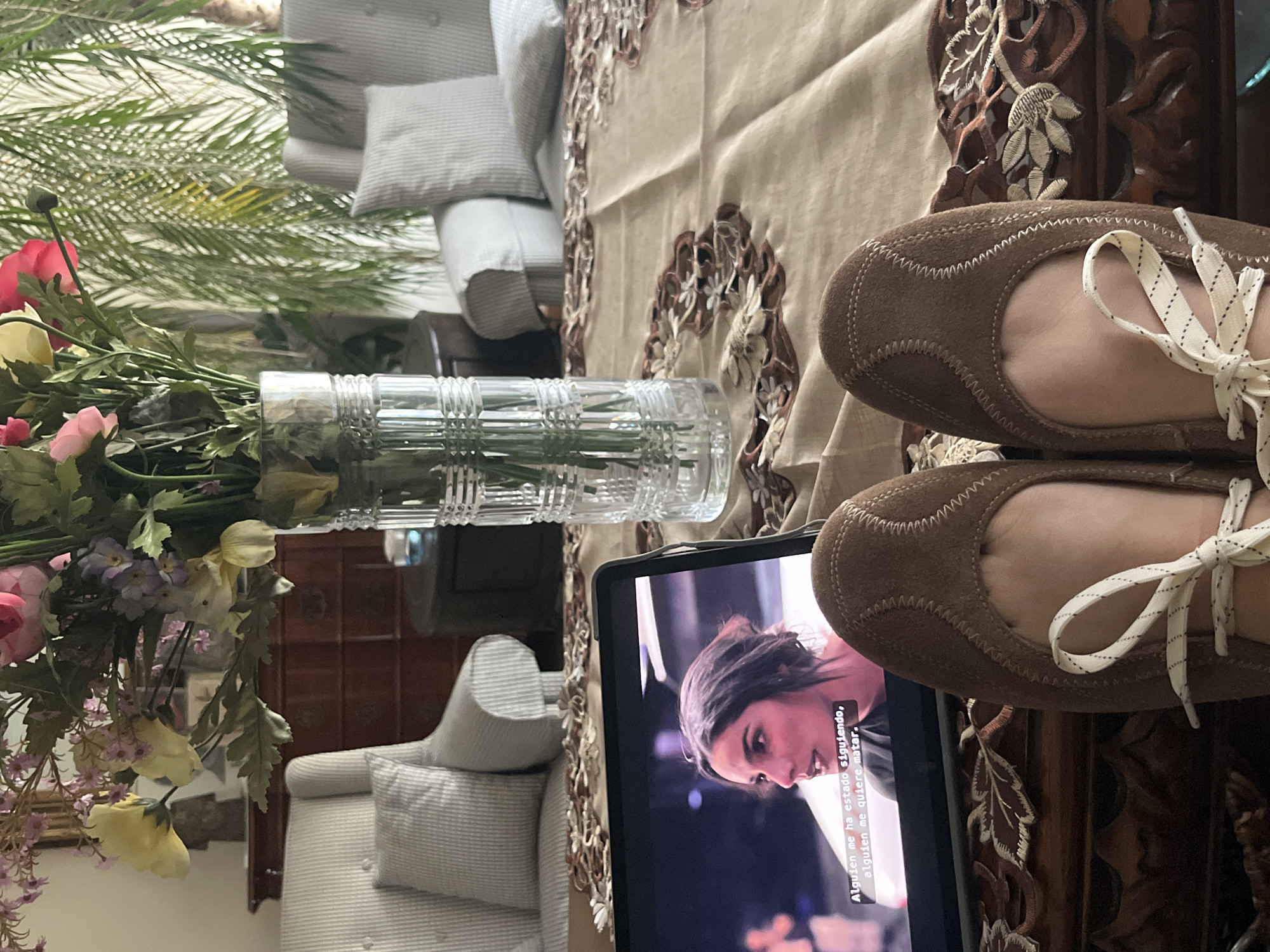 A low-angle, indoor shot depicts a person's feet wearing brown suede shoes with white laces, resting near a tablet displaying a woman's face. The tablet sits on a wooden table with intricate carvings and a patterned beige tablecloth. A clear glass vase filled with flowers is to the left, next to a gray and white striped chair. In the background, more furniture, including a cabinet and a sofa, is visible. Subtitles in Spanish appear on the tablet screen, suggesting that the video being watched is in Spanish. Based on the style of furniture and decor, the location might be a residential area in Mexico City, Mexico.