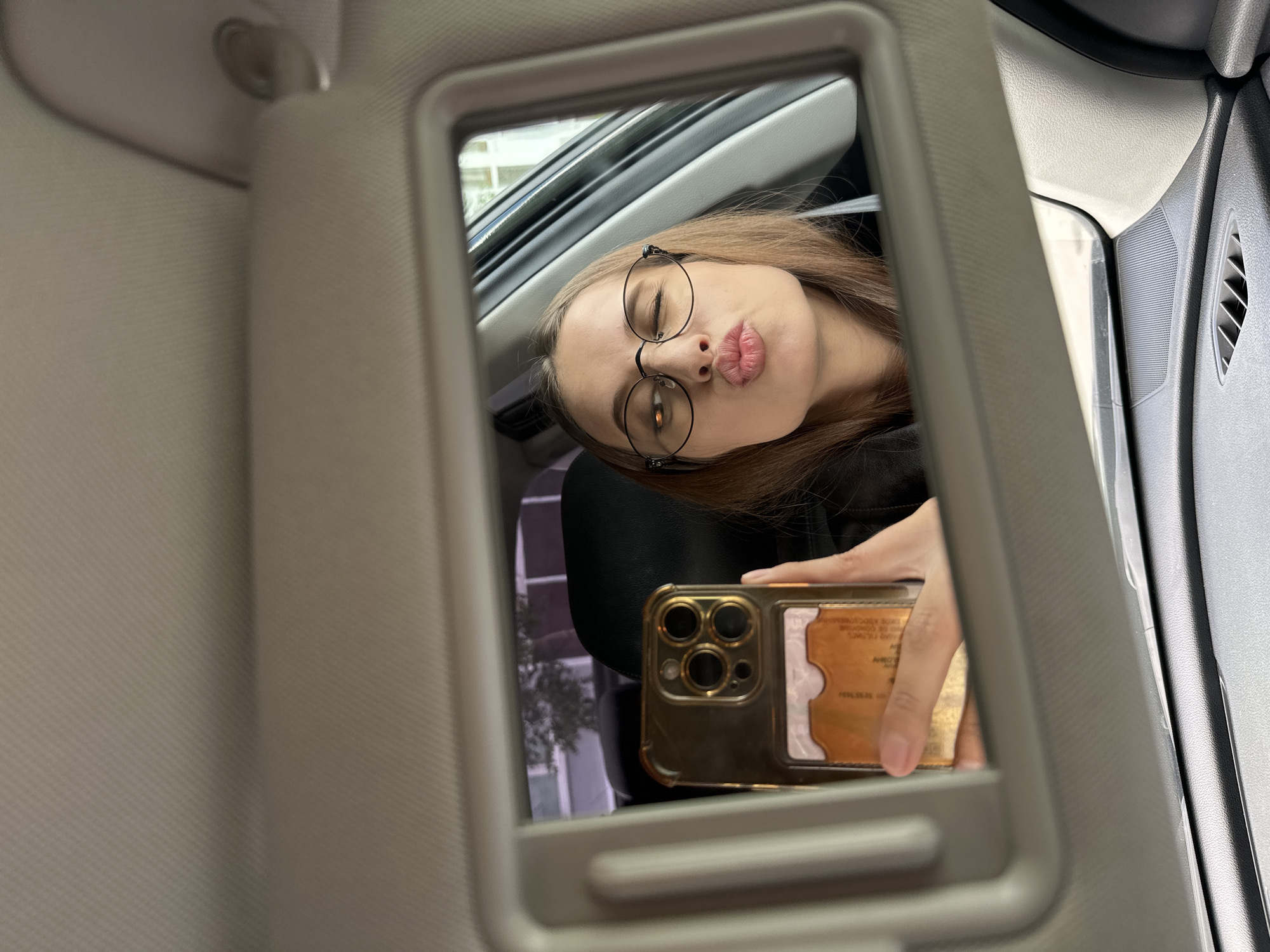 A female individual is depicted taking a self-portrait (selfie) reflected in the vanity mirror of a car. Her face, framed by dark-rimmed eyeglasses, shows a winking left eye and puckered lips. Her hair is light brown. She holds a smartphone, gold in color with a prominent multi-camera system (three lenses visible plus a flash/sensor array), encased in a transparent, gold-tinted cover. An attached cardholder accessory on the back contains a single light-brown colored card. The interior of the vehicle, located in Yekaterinburg, Russia, features a light beige/grey textured headliner and sun visor, with the vanity mirror being a component of the latter. Below the mirror, a dark grey textured dashboard with a rectangular air vent is visible, along with a light grey A-pillar or door frame on the right. Through the car's rear side window, reflected within the mirror, a light-colored multi-story building with dark windows and tree foliage is discernible, indicating an outdoor urban environment.