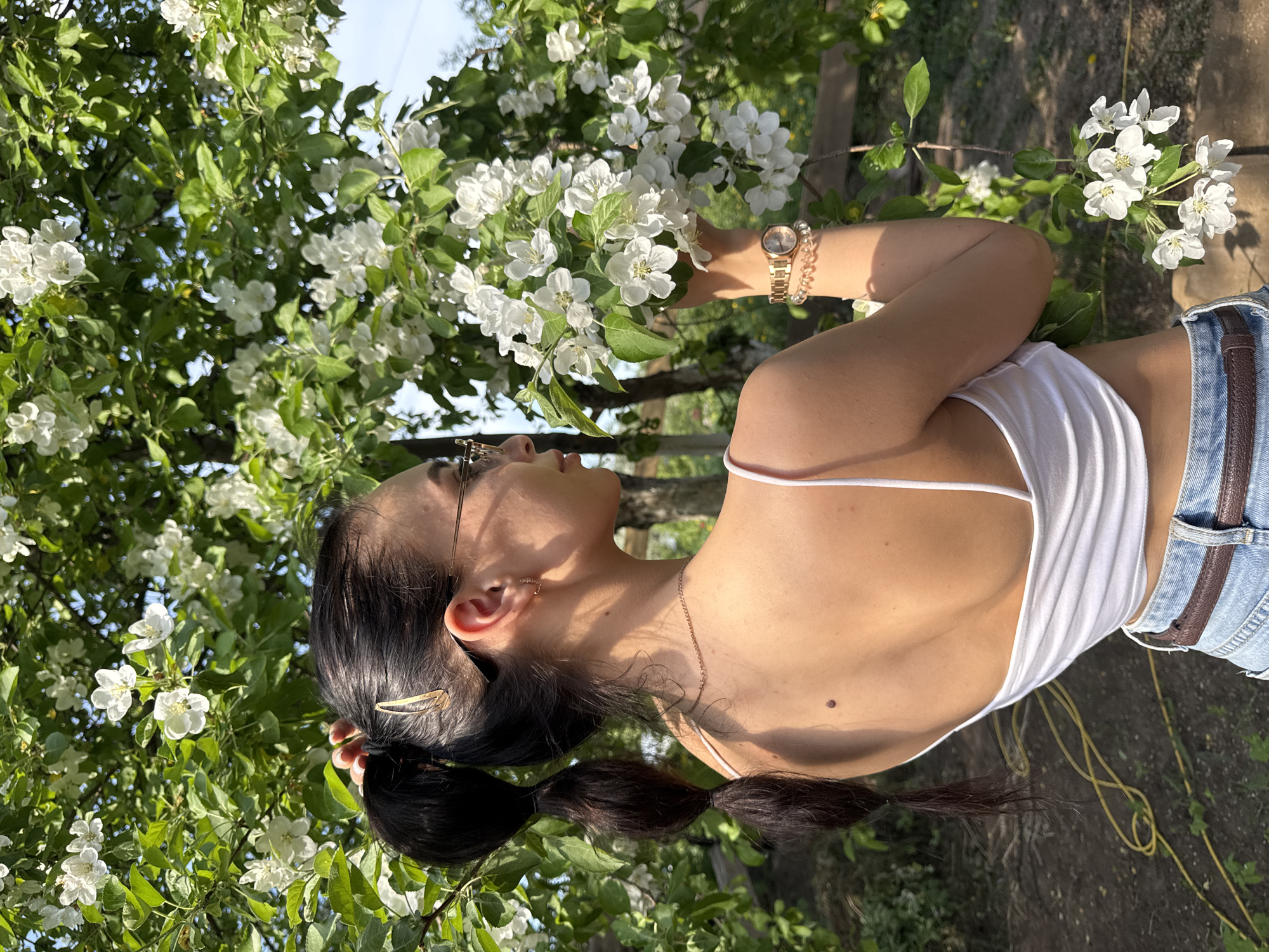A woman is seen in a natural outdoor setting, possibly in the city of Chaikovsky, Russia. The focal point is the woman wearing a white tank top and jeans with a brown belt. She has dark hair pulled back in a ponytail, and she is wearing glasses. She is standing amidst blooming white flowers, which could be part of a tree or shrub. She is holding the flowers, appearing to admire them. She is wearing a watch and bracelet on her wrist. The lighting suggests it is daytime, and the background indicates a green, possibly garden-like environment.