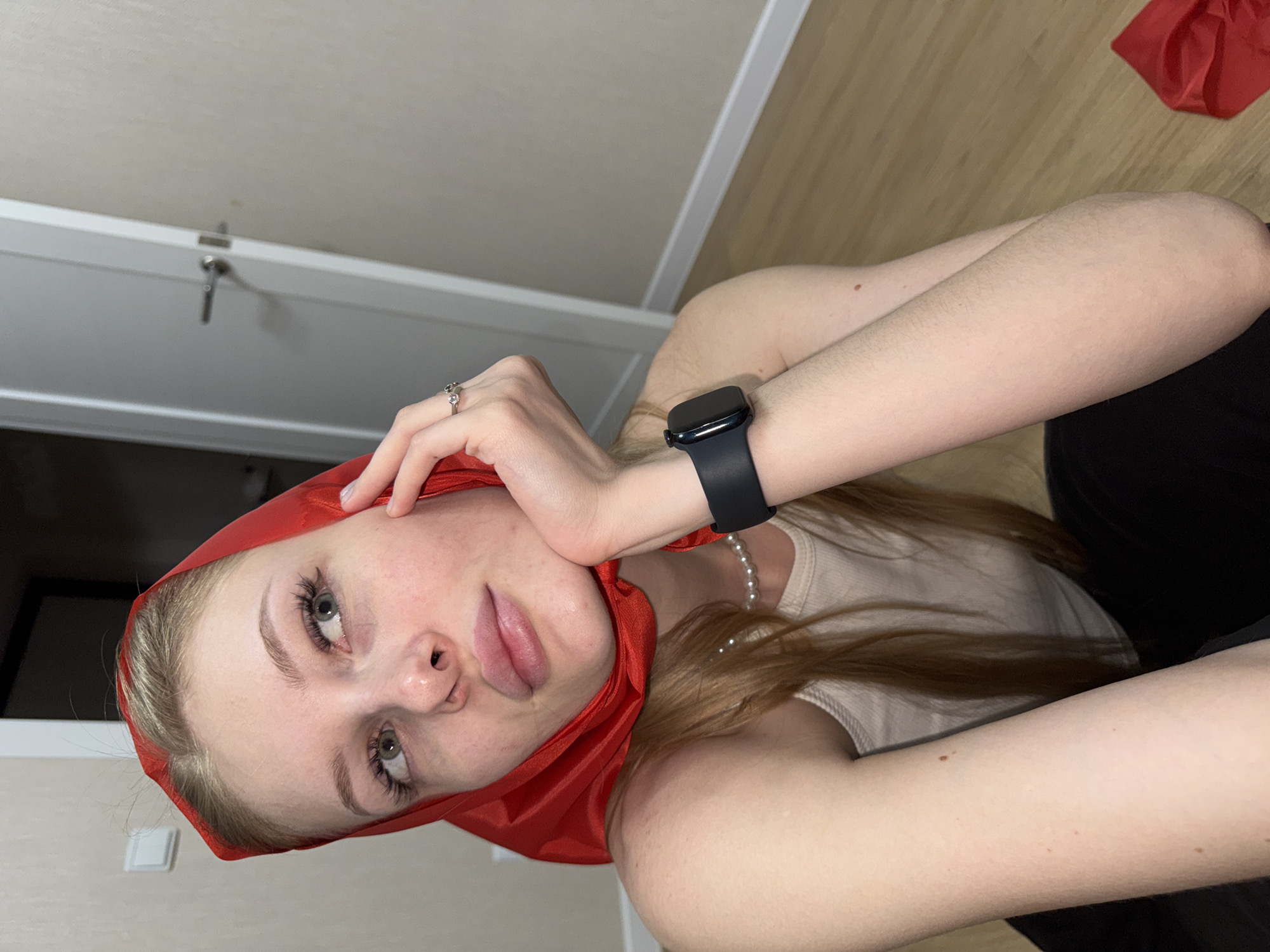 A young woman with long blonde hair and green eyes is sitting on the floor in a room. She is wearing a red headscarf, a beige tank top, and black pants. She has a pearl necklace and a smartwatch on her left wrist, and a ring on her right hand. Her right hand is resting on her chin, and she is looking up and to the side. The room has light-colored walls and a wooden floor. A white door is visible in the background. A red scarf is lying on the floor to her right. The scene appears to be indoors, in a residential or domestic setting. There are no indications to suggest the location is in Perm, Russia.
