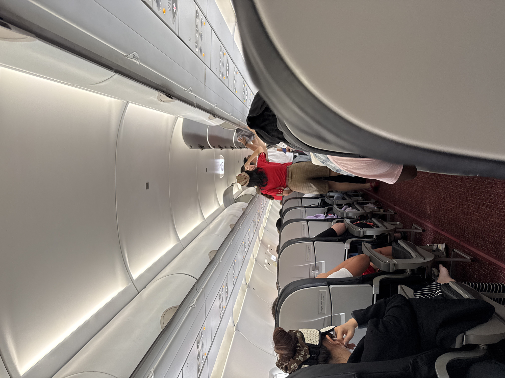 The image shows the interior of a commercial airplane cabin. Passengers are seated, with some standing near the overhead compartments. The cabin has overhead bins, passenger service units with reading lights and air vents, and seats arranged in rows. The carpet is red. The scene is not explicitly linked to Đà Nẵng, Vietnam.