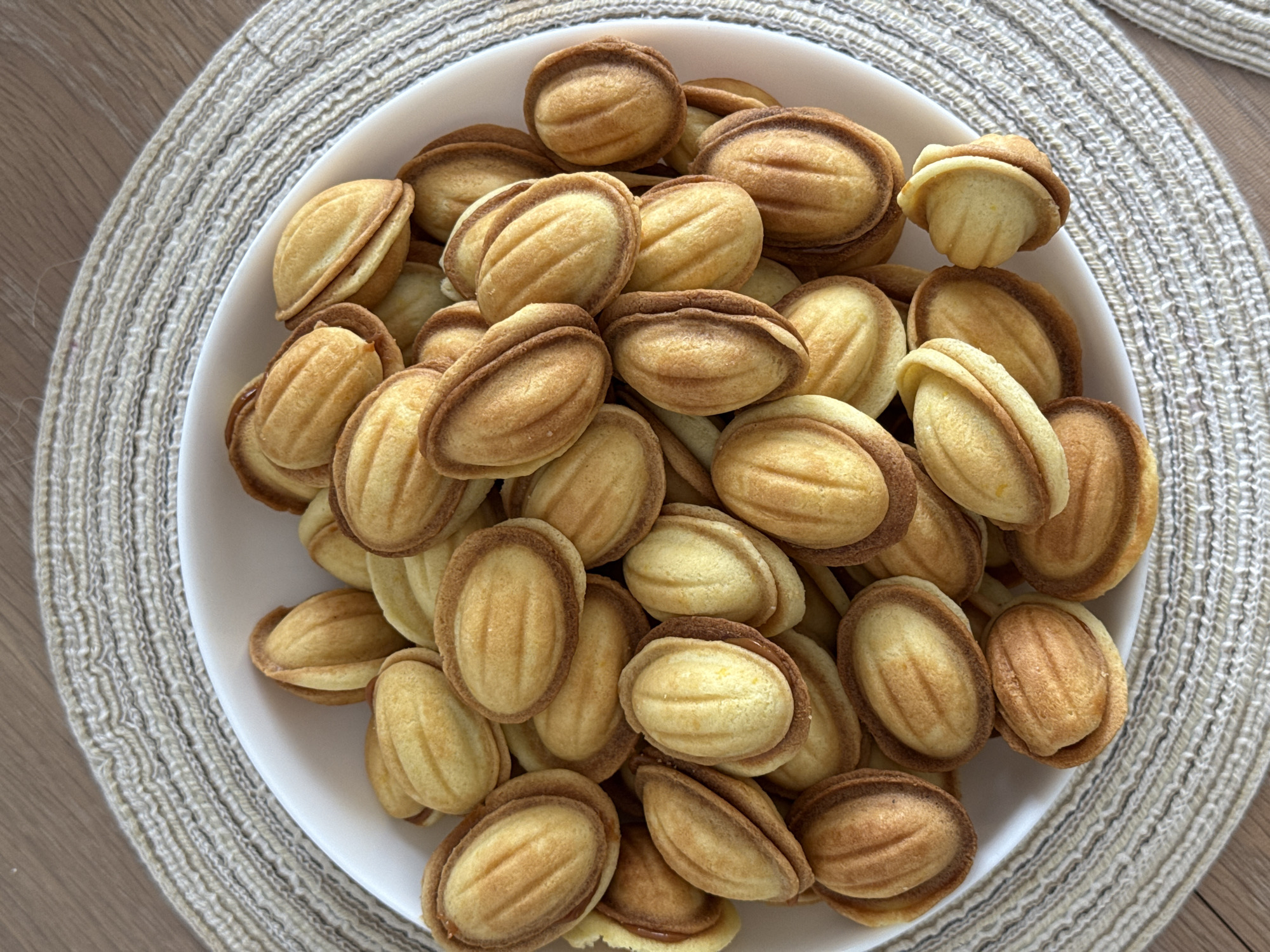 A top-down view captures a white, round plate overflowing with numerous small, oval-shaped cookies. The cookies, commonly known as Oreshki, feature a light golden-brown color with slightly darker, ridged edges, resembling small nuts or shells. Many of the cookies show two halves joined together, with some hinting at a creamy, light brown filling peeking out between the shells. The cookies are densely packed in the plate, creating a mound.
The white plate sits on a light-colored, round placemat with a woven texture, showing distinct threads in a slightly irregular pattern. Beyond the placemat, a light brown wooden surface with a visible grain is partially visible, providing a simple and natural background. The lighting is even, suggesting an indoor setting. There are no people present, no discernible activity beyond the display of food, and no visible text. The overall scene suggests a casual home environment, with the cookies presented for enjoyment.