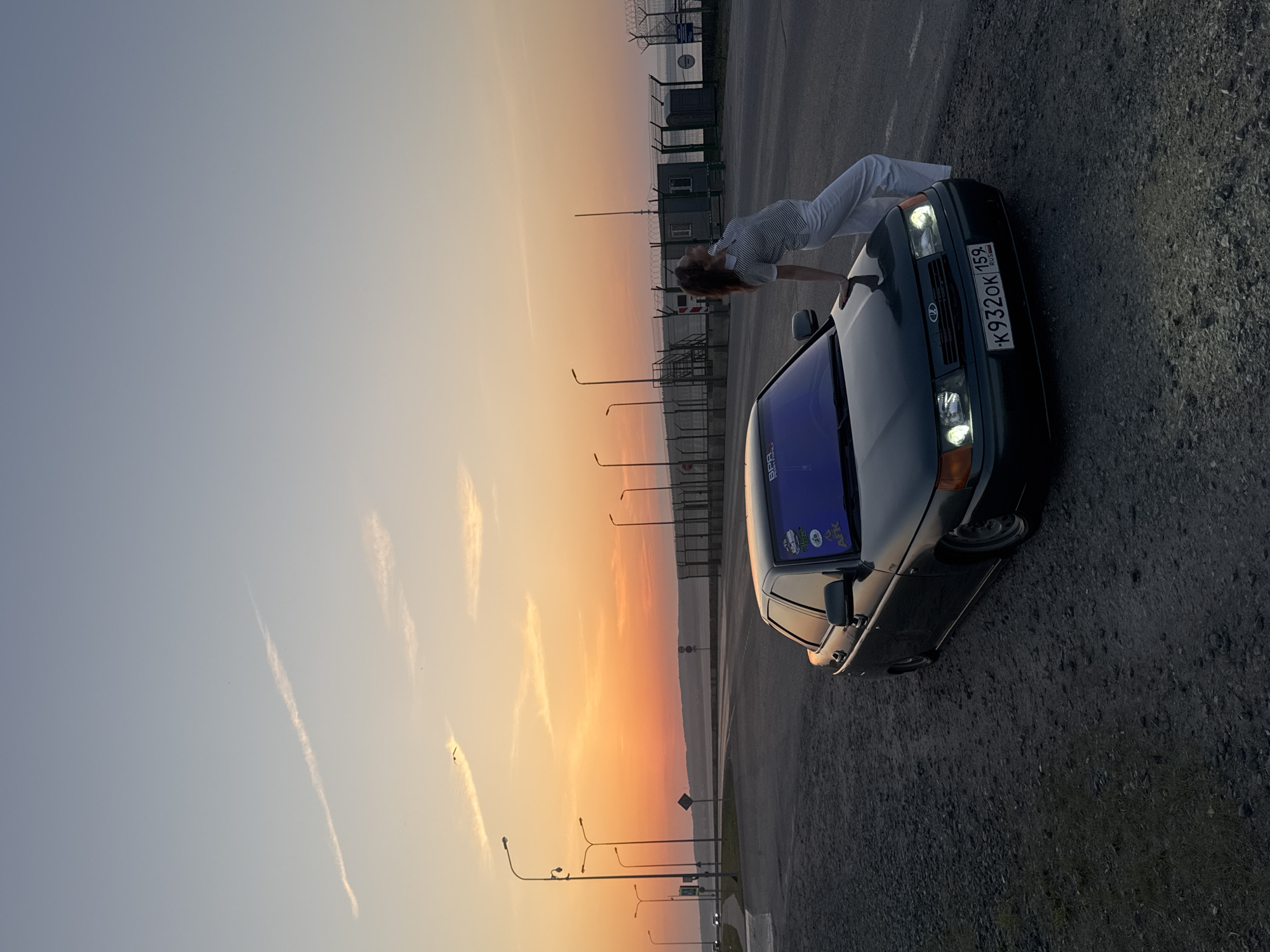 The image shows a woman in a white striped shirt and white pants leaning over a dark-colored car, possibly a Lada, with the license plate "K932OK159 RUS" visible. The car's headlights are on. The setting appears to be a roadside near a fenced-off area and streetlights, with a gradient sky suggesting either sunrise or sunset. The location is likely Chaikovsky, Russia, based on the license plate designation (159). The ground near the car is a mix of asphalt and gravel. The woman's face is obscured by her leaning posture.