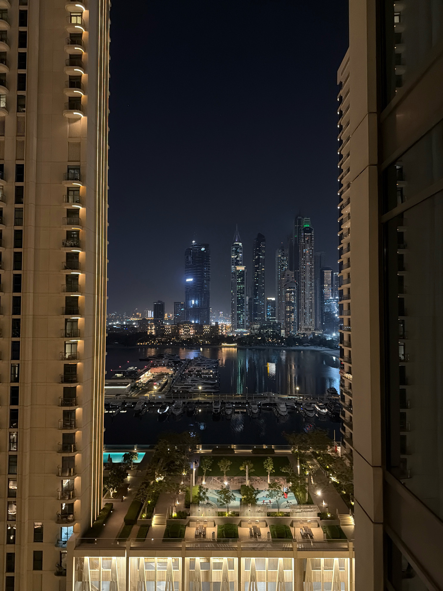The image depicts a nighttime view of the Dubai Marina in Dubai, United Arab Emirates. Two modern residential buildings frame the scene, with the one on the left illuminated by vertical golden lights along its facade, highlighting its balconies and windows. The building on the right, viewed from its side, shows similar balconies and windows.
Between the buildings, the Dubai Marina is visible, filled with numerous yachts docked in the water. The water reflects the lights of the surrounding city. Beyond the marina, a cluster of distinctive Dubai skyscrapers dominates the skyline, illuminated against the dark night sky. The most prominent skyscraper on the right side of the skyline is tall and sleek, with a rounded top. Other skyscrapers of varying heights and designs are also visible.
In the foreground, below the marina and between the two buildings, is a landscaped outdoor area. This area includes a swimming pool, trees, manicured bushes, outdoor seating, and walking paths, all subtly lit by artificial lighting, suggesting a residential complex or hotel amenity. The overall impression is one of urban luxury and a vibrant cityscape at night.