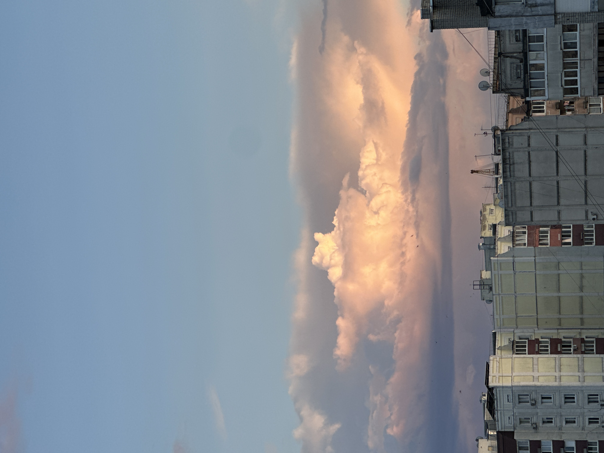 The image shows a view of apartment buildings against a sky filled with clouds. The clouds are illuminated with warm, pink and orange hues, suggesting either sunrise or sunset. The buildings are multi-story structures with numerous windows and visible architectural details. Some buildings appear to be made of concrete, while others have lighter colored facades. Several satellite dishes and antennas are visible on the rooftops. A few small birds are captured in flight against the cloudy sky. The scene is set in an urban environment, likely a residential area.