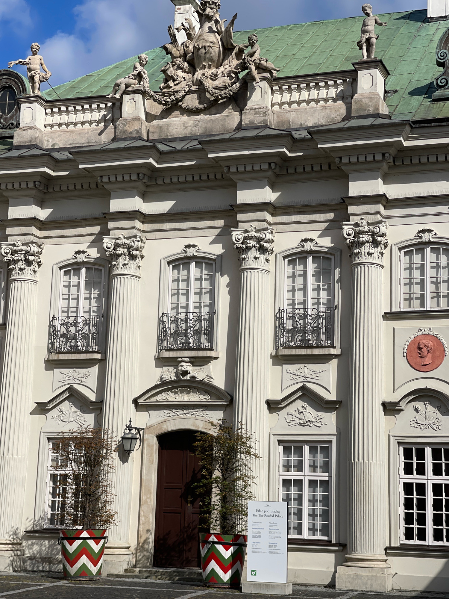 The image shows the exterior of the Tin-Roofed Palace (Pałac pod Blachą) in Warsaw, Poland. The palace is a light-colored, Baroque-style building with a green roof. Sculptural elements adorn the facade, including figures on the roofline and ornate capitals on the columns. There are several tall, white-framed windows with black wrought-iron balconies. A dark brown wooden door is visible to the right of center. In front of the door, two large planters with a red, white, and green chevron pattern contain small trees. A white information sign is placed in front of the left planter. The sky above is blue with some white clouds.