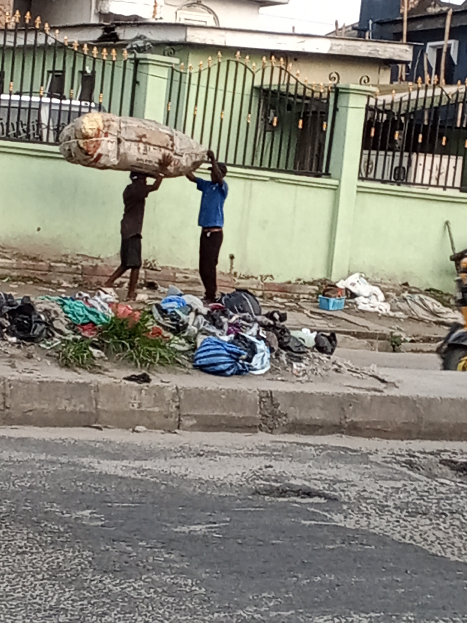 The image captures an outdoor street scene, likely in an urban or semi-urban area of Ado, Nigeria. Two individuals, appearing to be men or older boys, are actively engaged in transporting a large, oblong bundle. This substantial package, wrapped in light-colored sackcloth or tarpaulin and secured with rope, is balanced on the head of the person in the rear, who is dressed in dark shorts and a dark top. The person in front, wearing a blue t-shirt and dark trousers, assists by supporting the bundle with their hands. This method of carrying heavy loads on the head is a common sight in many parts of Nigeria and West Africa.
The setting includes a paved road in the immediate foreground, separated by a concrete curb from a dirt and grass shoulder. Along this shoulder, numerous piles of assorted textiles, clothing, and miscellaneous items are scattered, suggesting either an informal street market, items being sorted, or discarded goods. A blue plastic basin is visible further back amidst the clutter on the right side of the frame.
In the background, a light green wall topped by a black metal fence with ornate golden accents forms the boundary of a property. Parts of buildings, including a darker structure, are visible behind the fence. The lighting indicates it is daytime, possibly under an overcast sky or during the softer light of early morning or late afternoon, as strong shadows are absent. The road surface appears worn in places. No clear or legible text is discernible on any objects within the frame.