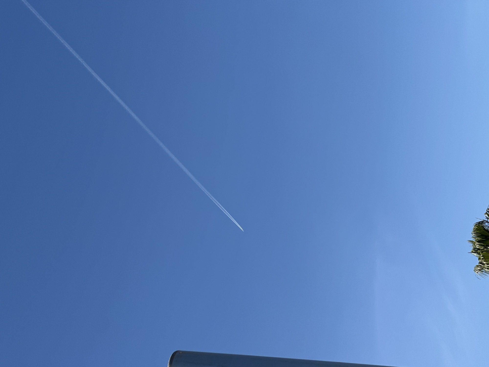 The image shows a clear blue sky. A contrail from an airplane stretches diagonally across the sky from the upper left towards the center. In the lower center and left is a silver metal pole. In the lower right corner is a palm tree. It is not possible to determine the specific location in the city of Málaga, Spain based on this image alone.