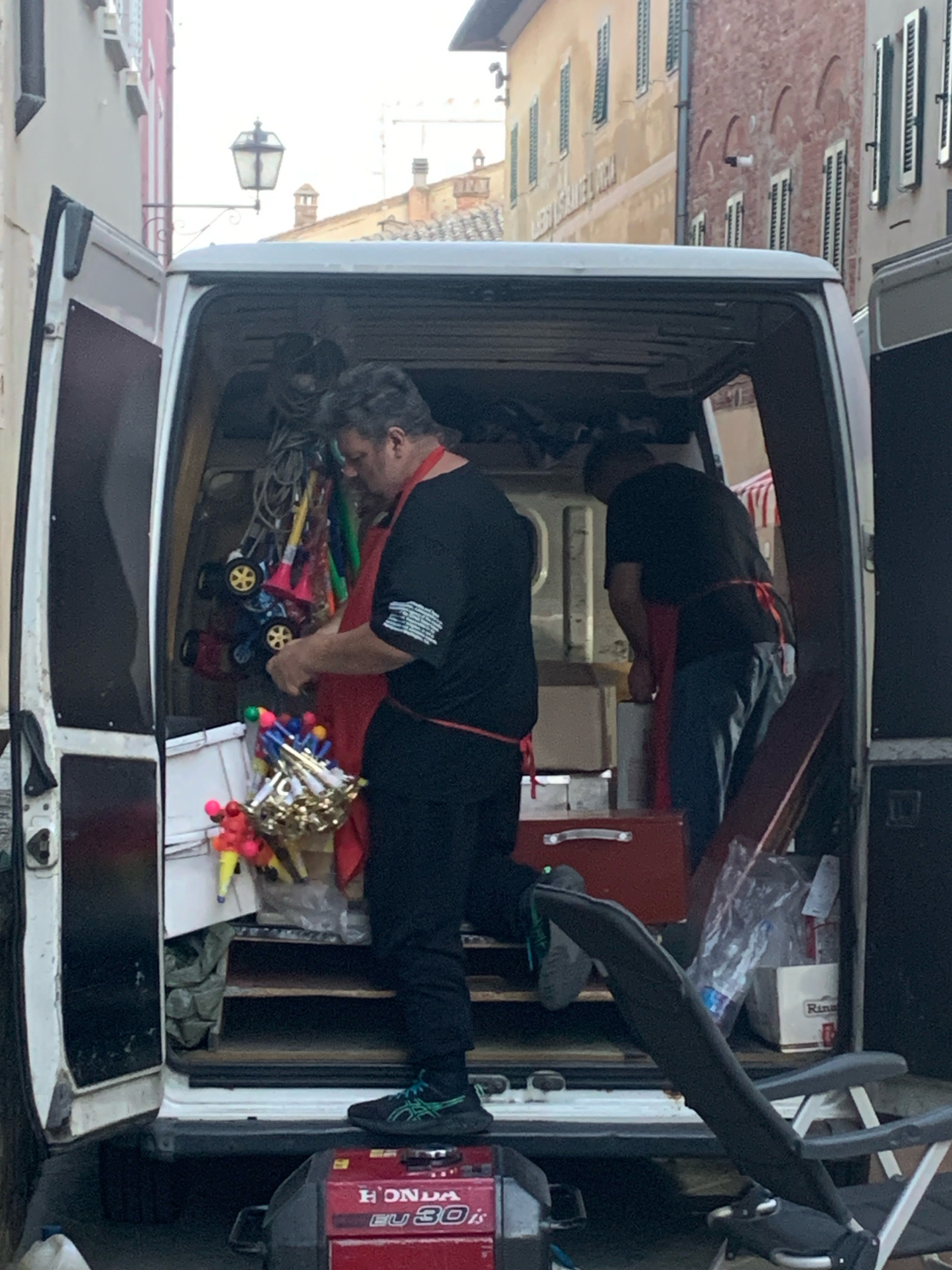 In San Quirico d'Orcia, Italy, a man wearing a black t-shirt and an orange apron stands inside the open rear of a white van. He is interacting with several colorful party horns and noise makers, some of which are hanging from the van's interior and others are in a bin on the van floor. Another person is partially visible on the left, also wearing an orange apron. The van is packed with various items, including boxes, bags, and what appears to be a portable generator in front of the vehicle. The scene suggests preparation or packing of items for an event.