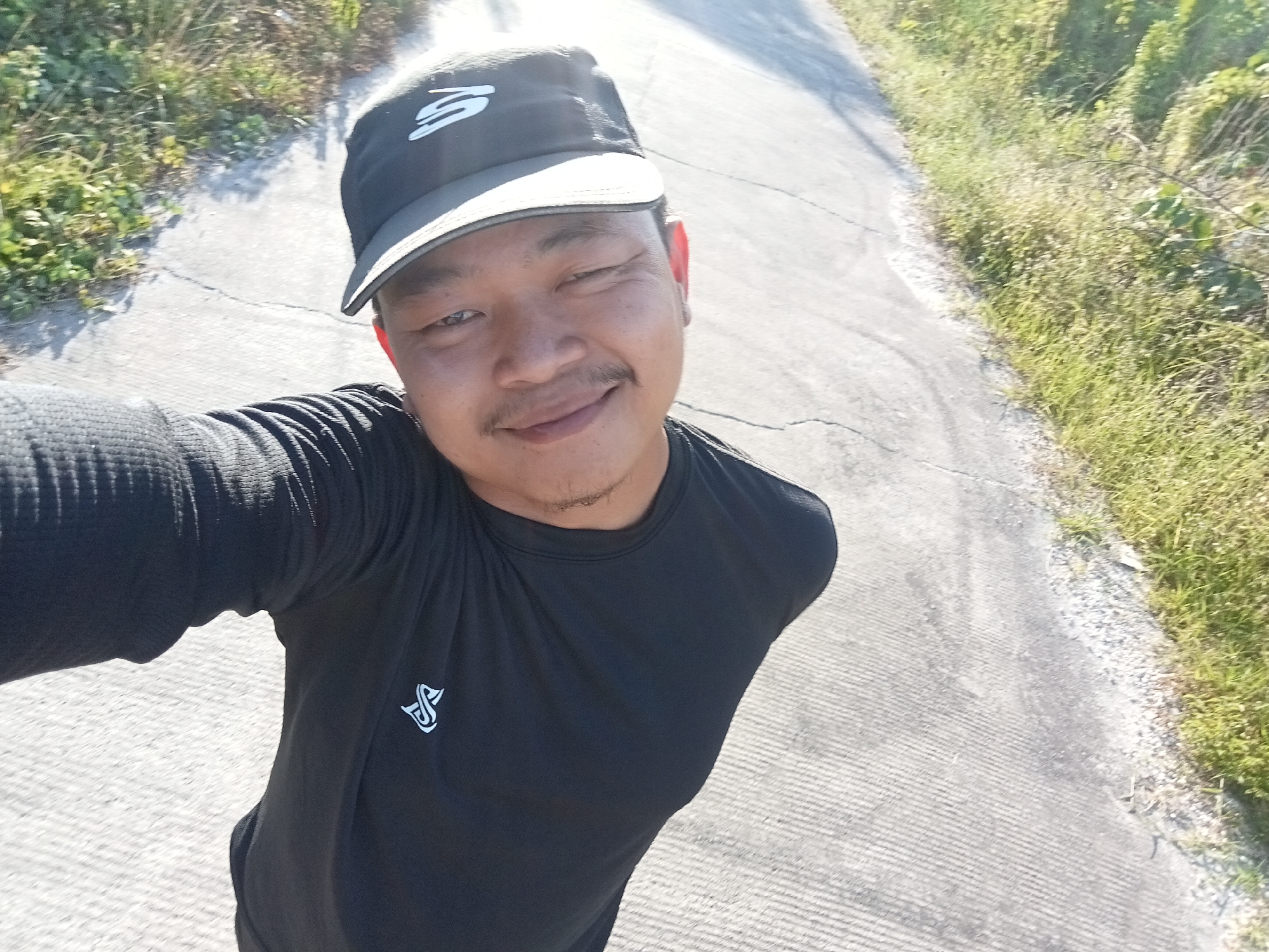 A man wearing a black t-shirt and a black baseball cap with a white logo is standing on a paved road in Kualu, Indonesia. The road has faint white lines and is bordered by dry grass and vegetation on the right side. The man is looking at the camera with a slight smile and his eyes are partially closed. The background shows more of the road and vegetation. The sun appears to be shining, casting shadows on the road.