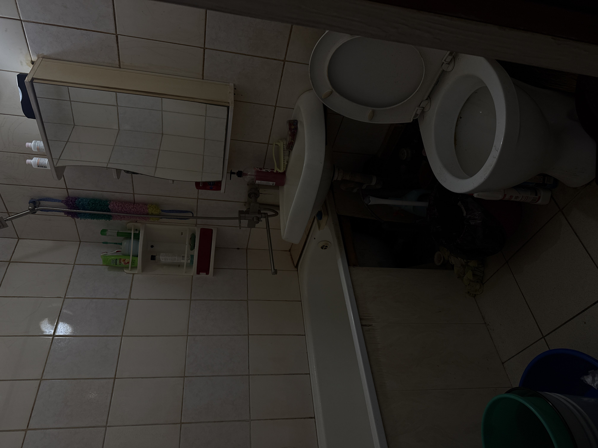 A bathroom in Baikonur, Kazakhstan, contains a white bathtub with a faucet, a white sink with a faucet, a toilet, and a medicine cabinet. The bathroom walls are tiled with beige square tiles. The floor is also tiled with beige square tiles. There are various items on the shelves and around the sink, including bottles of soap and shampoo, a toothbrush, a sponge, and a trash can. A green bucket and a blue bucket are on the floor in the lower left corner of the image. The lighting in the bathroom is dim.