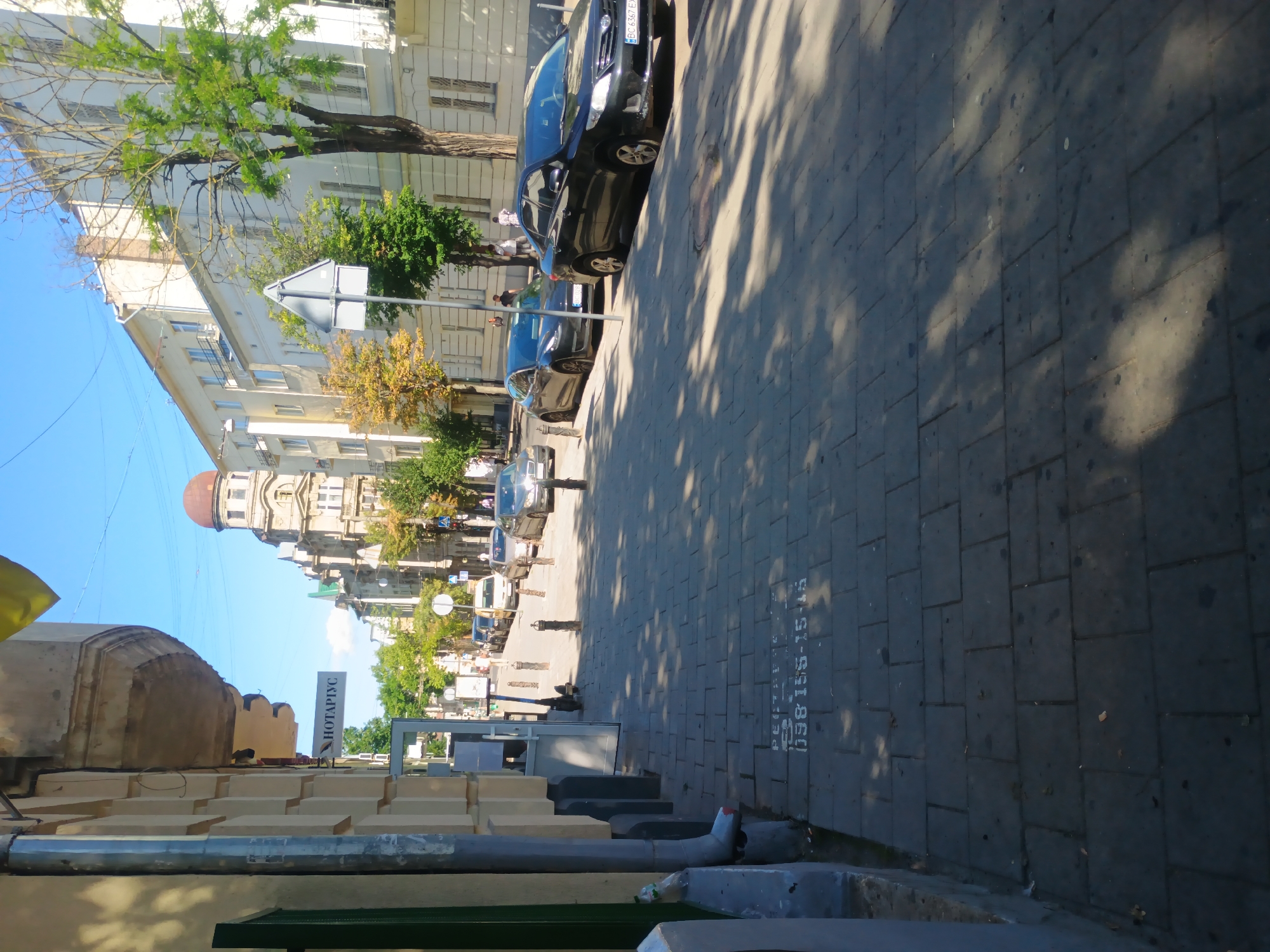 The image depicts a street scene in Lviv, Ukraine, on a clear, sunny day. The perspective is from a sidewalk looking down a street lined with buildings and parked cars.
On the left side of the frame, a building with a cream-colored facade is visible, featuring architectural details like cornices and a metal downspout. A sign above an entrance reads "НОТАРІУС" (Notary in Ukrainian). A green-painted doorway is also present.
The sidewalk is paved with gray rectangular stones, and the sunlight casts dappled shadows from trees. Some white spray-painted markings are visible on the sidewalk, including what appears to be a phone number: "098 155-15-15".
The street is lined with parked cars, including a black Toyota Corolla with a Ukrainian license plate "BC 6367 EX" and a dark gray Tesla. Further down the street, more cars and buildings are visible, with trees interspersed between them. In the background, a building with a domed roof is a prominent architectural feature.
Several street lamps and bollards are present along the sidewalk. The sky is a clear blue. There are a few pedestrians visible in the distance, walking on the sidewalk or crossing the street. One person appears to be riding a scooter. The overall impression is of a typical urban street scene in Lviv.