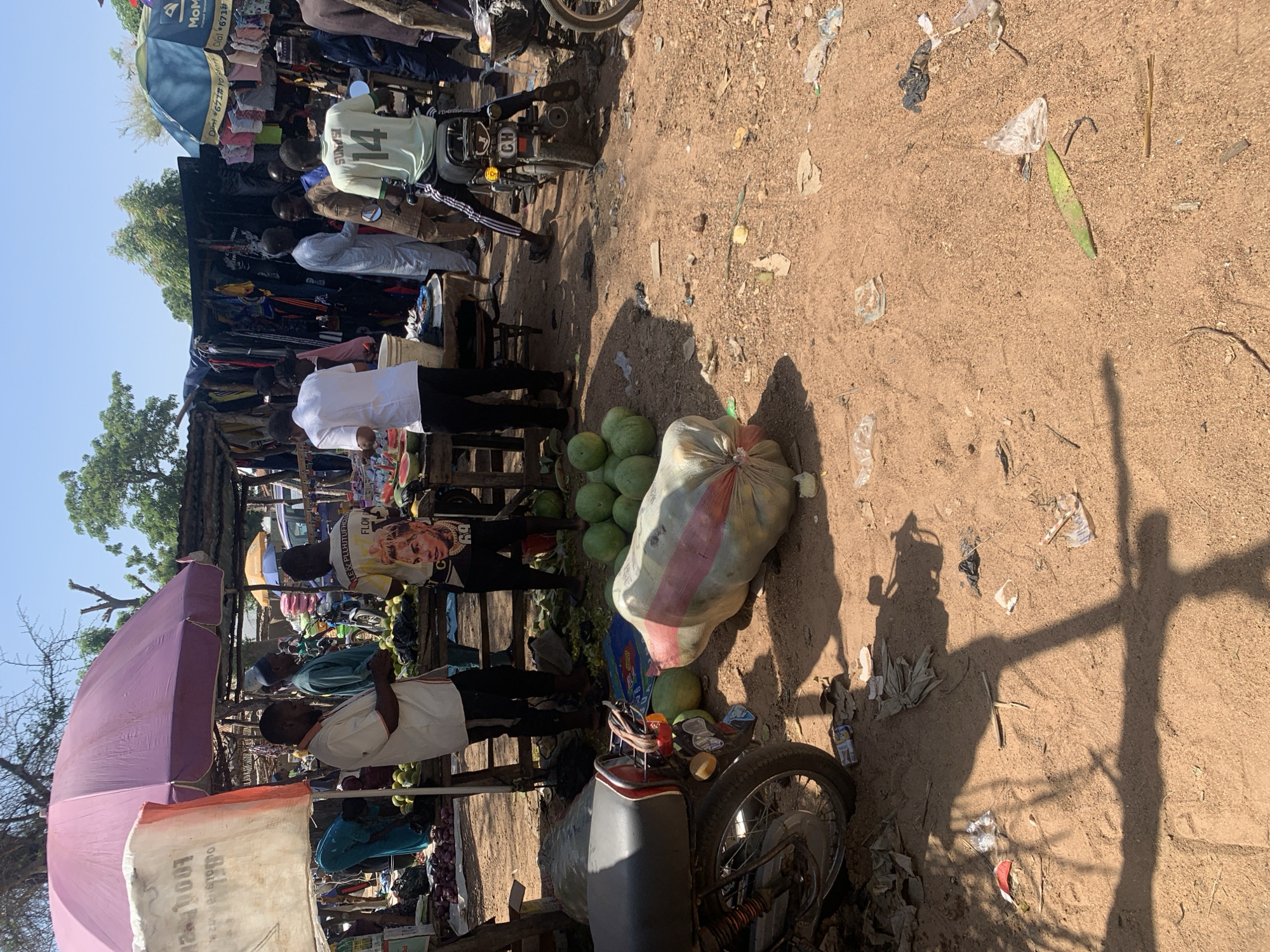 This media file captures a bustling outdoor market scene in Nigeria. The image is oriented vertically, presenting a ground-level view of a busy marketplace. Several individuals are visible, appearing to be vendors and shoppers. One person in the foreground is wearing a white shirt and black pants, and another to their left wears a white shirt with black sleeves. In the background, more people are present, some interacting with market stalls laden with goods.
The market is set up with wooden structures and colorful umbrellas, offering shade. Various items are displayed for sale, including clothing hanging from a stall under a blue umbrella with "MoM" text, and a pile of produce, likely watermelons, near a parked motorcycle. The ground is dusty, characteristic of many outdoor markets. Scattered on the ground are pieces of litter, including plastic bags and other debris, suggesting a well-used, unpaved area.
A motorcycle is parked prominently in the middle right of the frame. There's a large, roughly woven sack filled with what appears to be produce, possibly watermelons, next to it. The sunlight suggests it is daytime, likely late morning or afternoon, casting distinct shadows on the dusty ground. The overall atmosphere appears lively and active, typical of a Nigerian market.
Visible text includes "MoM" on an umbrella, "Dial *671#" with instructions, "ALE & PLLHJTUFHCO" and "FLOW" on clothing, "SUNSI" on a shirt, and the number "14" on a light green shirt. The "CH" on the motorcycle's license plate is also visible.