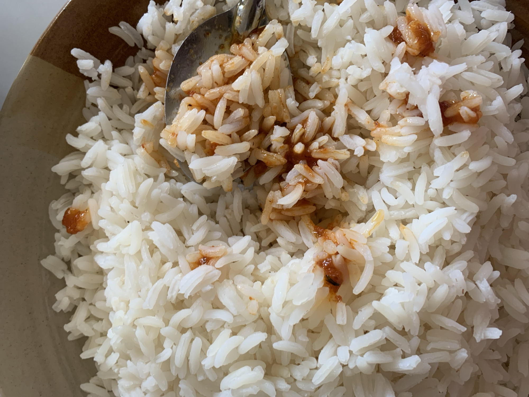 A close-up shot captures a bowl of white rice, primarily long-grain, being served or consumed. The main subject is a generous portion of fluffy, cooked rice filling most of the frame. A silver metal spoon is partially submerged in the rice, lifting a spoonful that is visibly mixed with a reddish-brown or orange-brown sauce. Traces of this same sauce are scattered throughout other sections of the rice in the bowl. The bowl itself is visible along the left and top edges, featuring an earthy-toned rim with a striped pattern, suggesting it is made of ceramic or pottery. No people are visible in the image. The scene focuses on the food item and the action of eating or serving. There are no visual cues within the image to indicate the time of day, weather conditions, or any specific emotions. No visible text is present. This meal is provided within the context of New Stadium, Nigeria, although the location itself is not depicted in the photograph.
