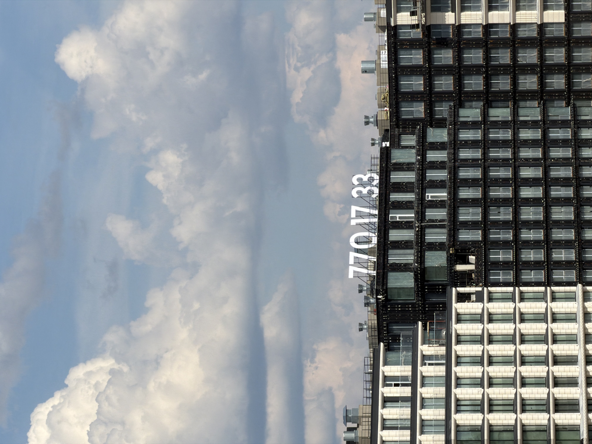 An aerial or high-vantage shot captures a contemporary urban landscape beneath a partly cloudy sky. The upper two-thirds of the image features a light blue sky interspersed with large, bright white cumulus clouds, accompanied by some thinner, darker cloud layers. The lower portion reveals the upper levels and rooftops of two distinct, modern architectural structures.
On the left, a multi-story building displays a light-toned, possibly beige or off-white, paneled facade with numerous uniformly sized rectangular windows, several of which incorporate small balconies with glass railings. Adjacent to it on the right is a multi-story building with a darker, seemingly black or dark gray, facade also characterized by a grid of rectangular windows. Some sections of this darker building show signs of ongoing construction or finishing work, with exposed elements.
Prominently situated on the flat rooftop of the darker building are large, white numbers: "770 17 33," which appear to be either illuminated or highly reflective. Both rooftops are equipped with various utilitarian elements, including ventilation units, pipes, and safety railings. There are no individuals visible within the scene. The distinctive "770 17 33" marking indicates the location as the **Nevskaya Ratusha complex, specifically associated with the "Stockholm" residential complex**, in Saint Petersburg, Russia.