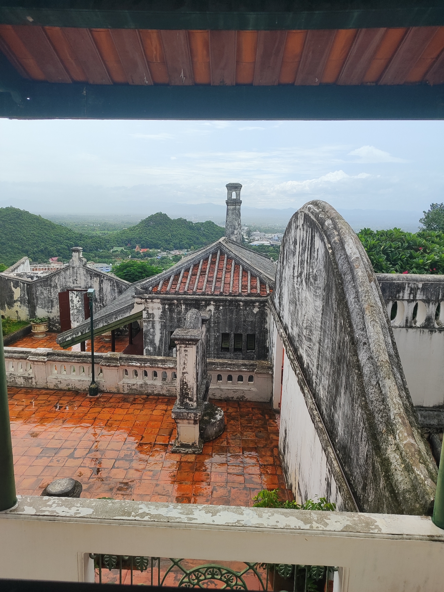 The image shows a view from a covered balcony overlooking a complex of old, weathered buildings with terracotta tile roofs. The foreground features the edge of the balcony railing and a wooden ceiling. The main focus is on a courtyard with wet, orange terracotta tiles, suggesting recent rain. Several architectural elements of the building are visible, including a prominent curved wall in the mid-right, white balustrades, and ornate stone pillars. In the background, a distant landscape of lush green hills and a hazy sky can be seen, with hints of a town or city nestled among the hills. A tall, square tower with an arched opening at the top is visible in the distance, and other buildings with red tile roofs are scattered across the landscape. The overall impression is one of an old, historic site, possibly a temple or palace, in a tropical or sub-tropical environment. This scene is characteristic of Phra Nakhon Khiri Historical Park, also known as Khao Wang, in Phetchaburi, Thailand, a former royal palace and temple complex situated atop a hill overlooking the city.