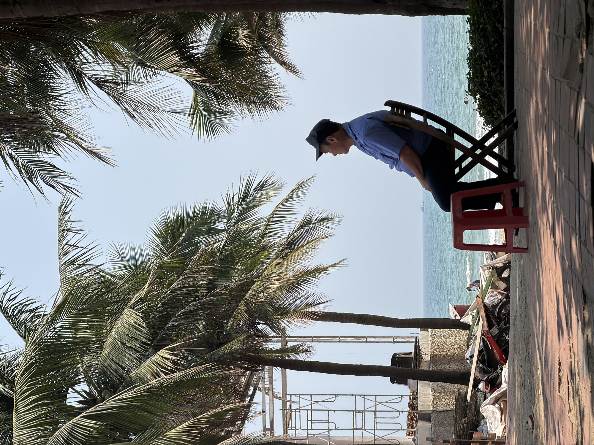 A man in a light blue shirt and dark pants sits on a red plastic stool, leaning forward in a cross-legged wooden chair, looking downward toward the ocean at Đà Nẵng, Vietnam. Palm trees frame the scene, and there is scaffolding in the background, along with assorted debris, including what appears to be a motorcycle. The sky is clear. The area is near a brick walkway or wall. The scene is rotated counter-clockwise in this composition.