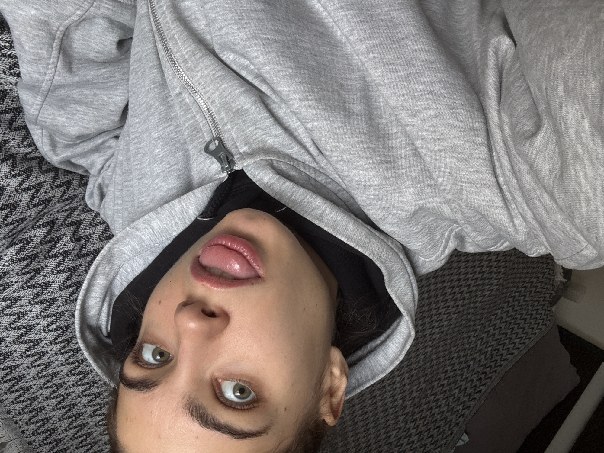 A woman is lying down, facing upward, wearing a gray hoodie partially zipped up to reveal a black garment underneath. She has fair skin, brown hair, and light-colored eyes. Her tongue is sticking out slightly. The background appears to be a gray and black patterned blanket or bedding. The image does not provide sufficient information to determine the location as Moscow, Russia, or any specific city.