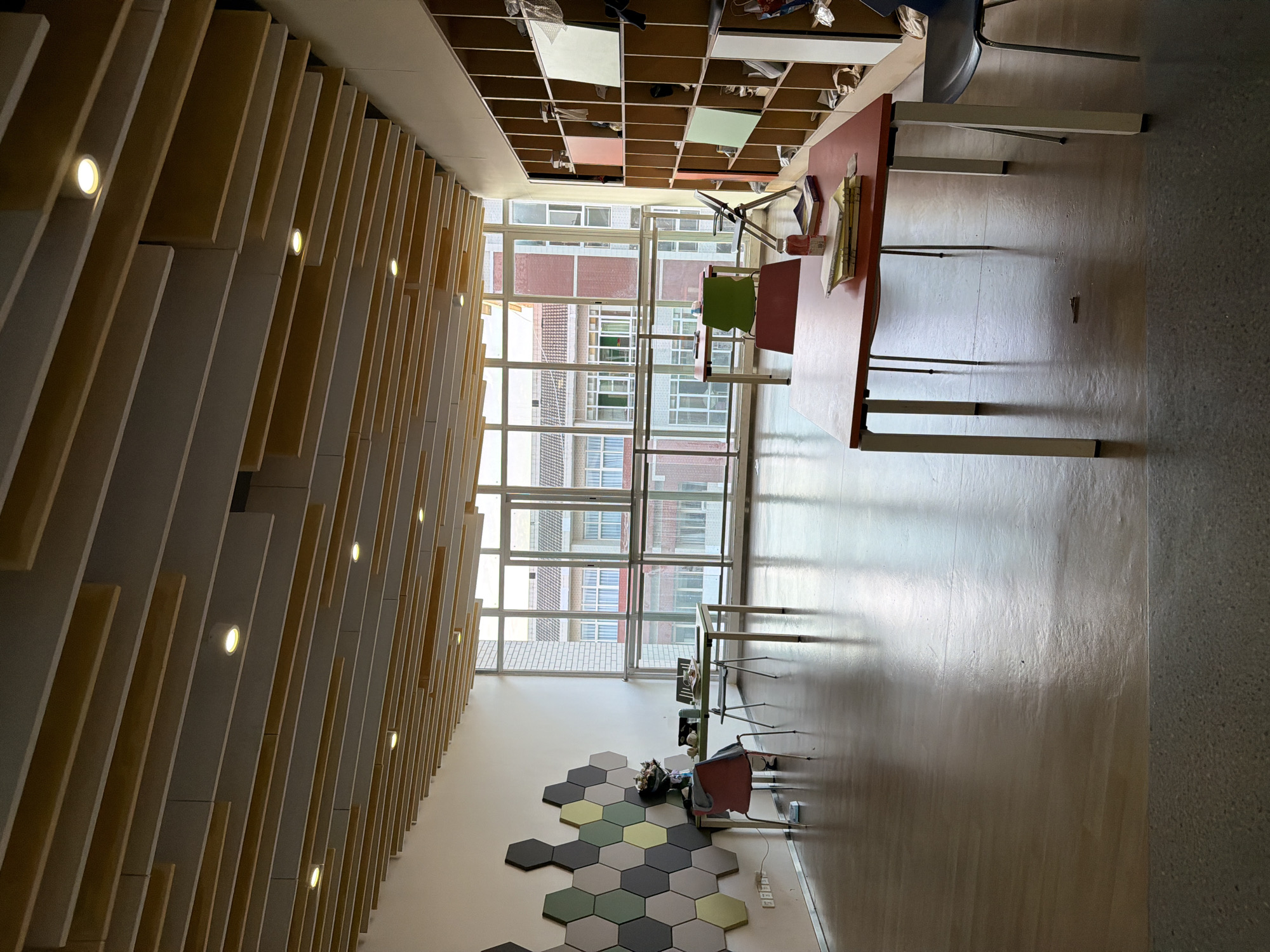 The photograph depicts an interior view, possibly within an educational or institutional building. The location is likely in the Chongqing High-tech Zone, China, considering the architectural style and cultural context implied by the scene.
The room is characterized by a modern design aesthetic. One wall features a decorative arrangement of hexagonal tiles in varying shades of gray, green, and yellow. Adjacent to this wall is a section of vertical wooden slats with embedded lights that extend along the ceiling. Large windows offer a view of an exterior environment that includes buildings.
The furnishings in the room consist of tables and chairs. The tables have red or white surfaces and metal legs, while the chairs are a mix of colors, including red and green. The tables are placed in different parts of the room, suggesting various areas for work or study. A storage structure with open compartments filled with items is located near the window. The floor is shiny and reflective.
There are no people visible in the photograph. The scene seems still and unoccupied at the time the picture was taken.