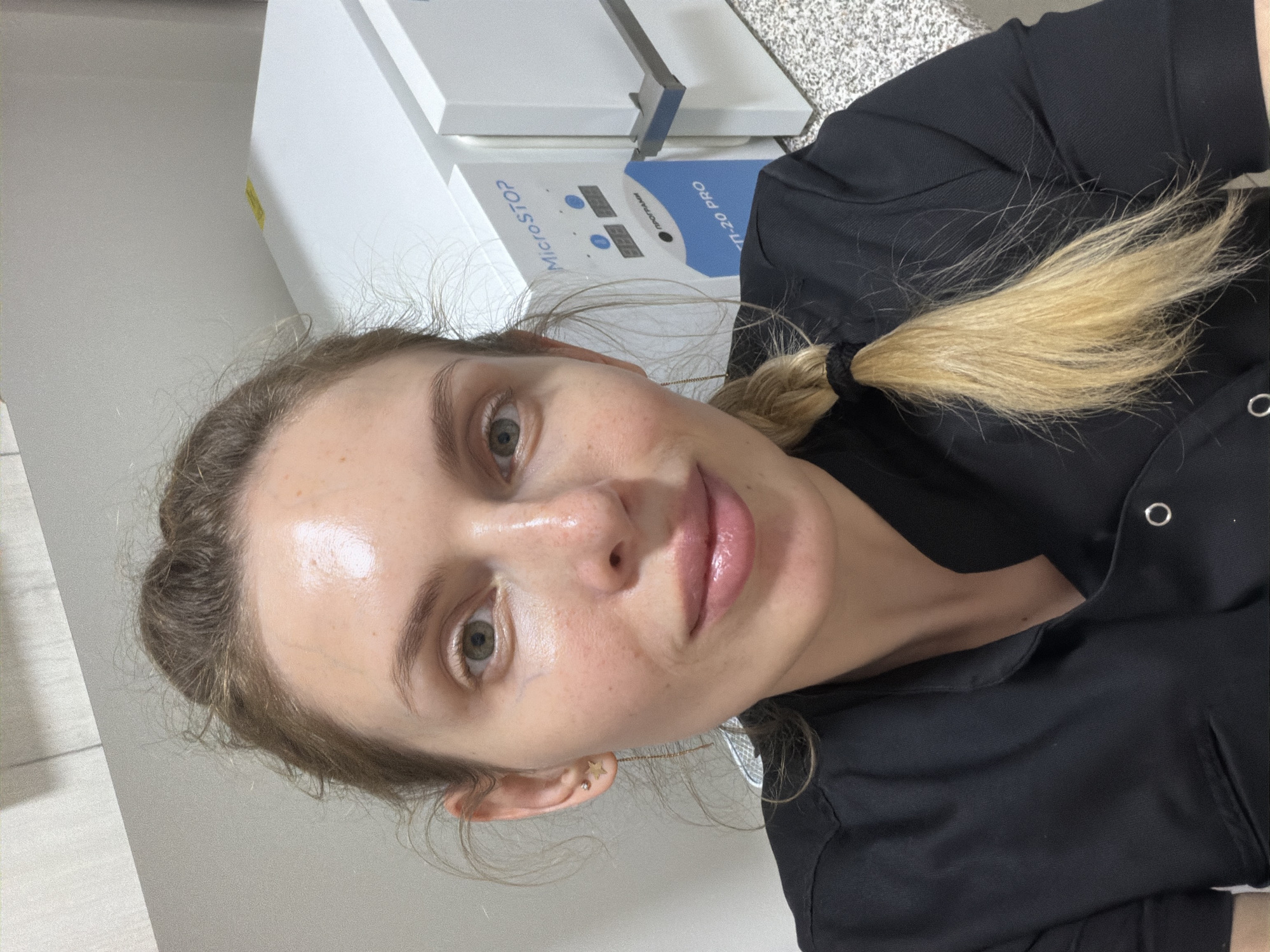 A young adult woman is captured in a close-up, head-and-shoulders selfie, looking directly at the camera with a neutral to slightly pleasant expression. Her light-colored eyes are open, and her lips appear slightly parted. She has light-blonde hair pulled back into a ponytail, with some loose strands framing her face. She wears a black top or uniform, visible around her shoulders and upper torso. In her left ear, two small stud earrings are visible: one is star-shaped, and the other is a simple round stud.
The setting appears to be an indoor professional environment, likely a clinic, salon, or lab, suggested by the equipment in the background. Behind her, a white and blue device is prominently featured, possibly a sterilizer or autoclave. Visible text on this device, which is oriented upside down in the image, includes "MICROSTOP", "ПРОГРАМЫ" (meaning "PROGRAMS" in Russian/Ukrainian), and "Т-20 PRO". There are also two small digital displays showing numerical readouts, possibly related to temperature or time. The background also shows light-colored walls and what appears to be a speckled or textured countertop surface. The overall lighting is even, suggesting an indoor setting without strong natural light cues. The woman's skin shows some natural freckling and a slight sheen, possibly from natural oils or a product. The location context for this scene is Budva, Montenegro.