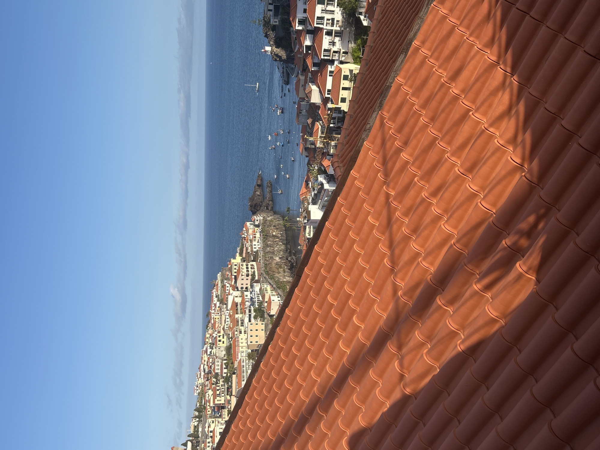 The image presents a view overlooking Câmara de Lobos, Portugal, from an elevated position, dominated by a terracotta tiled roof in the foreground. The ocean stretches towards the horizon, with a cluster of small boats visible near the coastline. The town is characterized by buildings with orange-tiled roofs, densely packed along the shoreline. The sky is predominantly clear blue. There are no people visible in the image. The scene is bathed in sunlight.