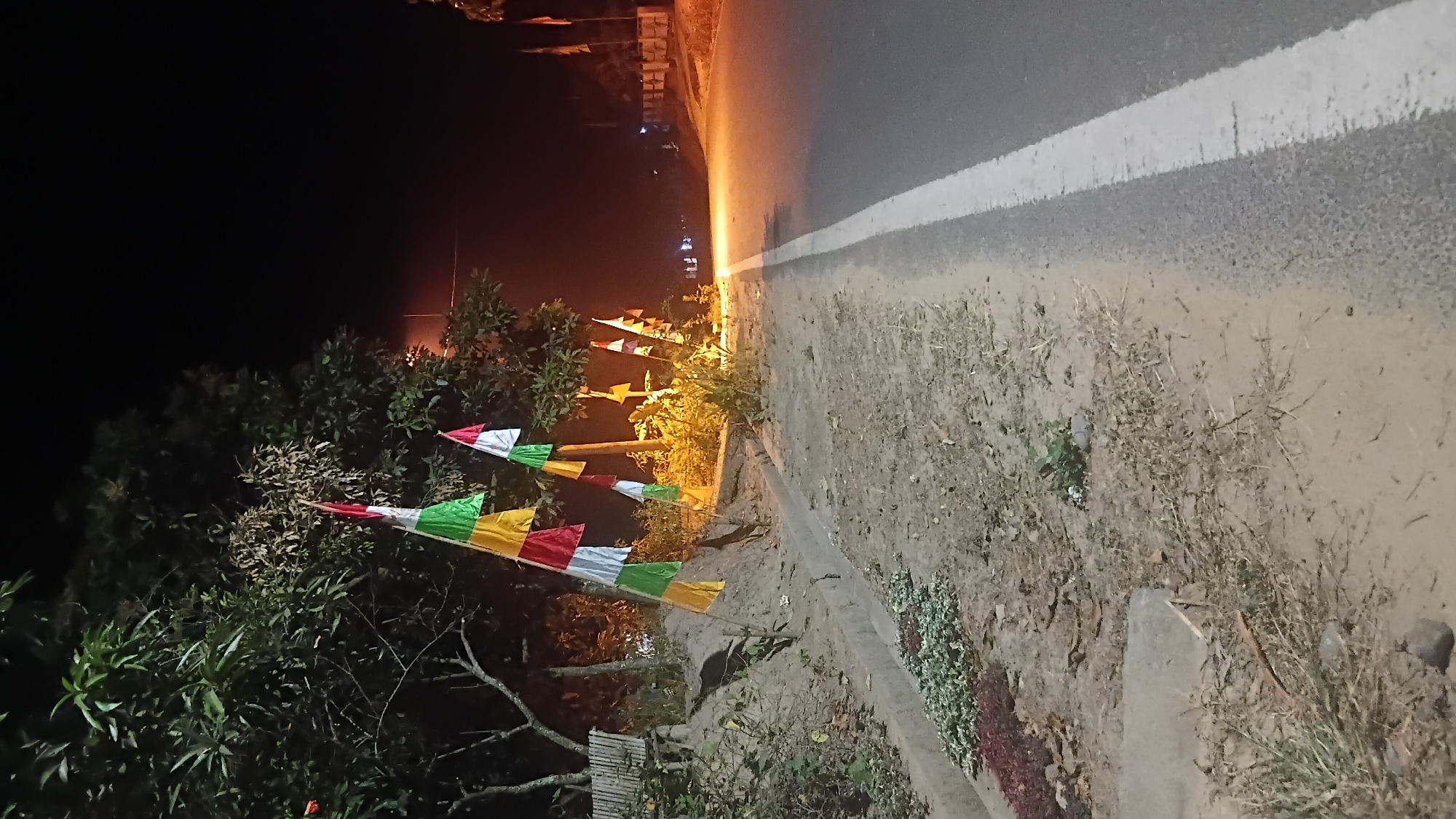 The image depicts a roadside scene at night in Prabumulih, Indonesia. The foreground shows a dirt and gravel shoulder bordering an asphalt road with a white lane marking. Vegetation, including dry grasses and some leafy plants, grows along the shoulder. A concrete ditch runs parallel to the road. In the background, several trees are visible, some with sparse leaves. Colorful pennant flags in red, white, green, and yellow are strung between poles and trees, suggesting a festive occasion. A distant street light illuminates the area. The overall impression is of a quiet, possibly rural or suburban, roadside at night.