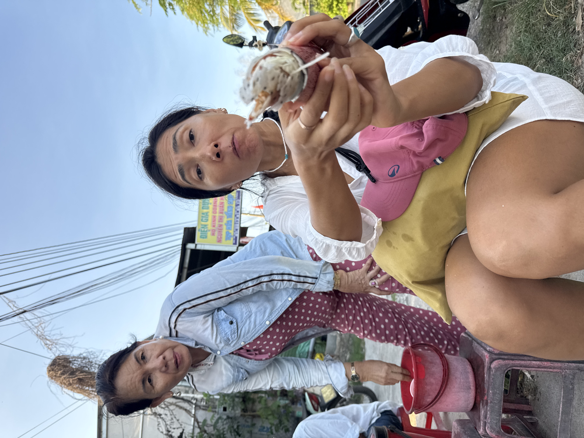 The image depicts two women outdoors, likely in Mũi Né, Vietnam, given the context of the street signage. The woman in the foreground is holding a food item, possibly a dessert or snack, to her mouth, suggesting she is eating it. She is wearing a white top and white shorts, with a yellow pillow and a pink cap resting on her lap. Her hands are adorned with rings. The woman in the background is wearing a denim shirt over a polka-dot dress and is looking towards the foreground woman. A red bucket sits on a small stool beside her. The environment includes a street with overhead wires and a clear sky. A motorcycle is partially visible in the background, suggesting a common mode of transportation in the area. The scene appears to be a casual outdoor setting, possibly a street food stall or a roadside stop.