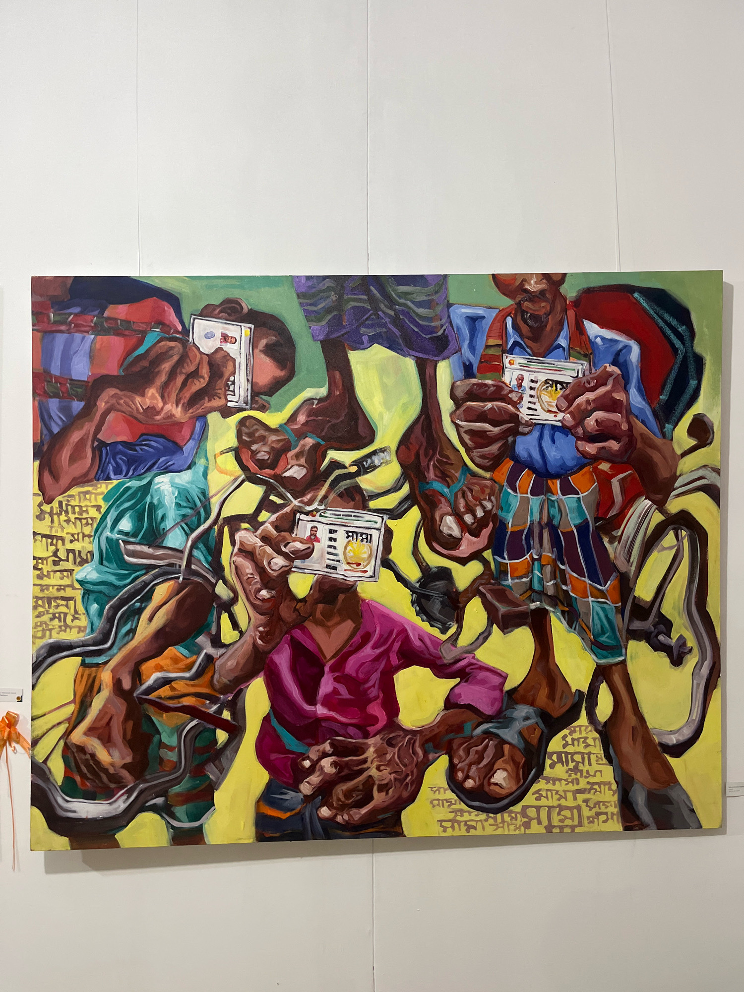 A painting depicts several individuals, each holding up an identification card. The figures are rendered in a distorted, fragmented style, with their limbs overlapping and their bodies contorted. The background is a vibrant yellow, with Bengali script inscribed in a repetitive pattern. The overall impression is chaotic and surreal. The artwork is displayed on a white wall, presumably in an art gallery in Dhaka, Bangladesh.