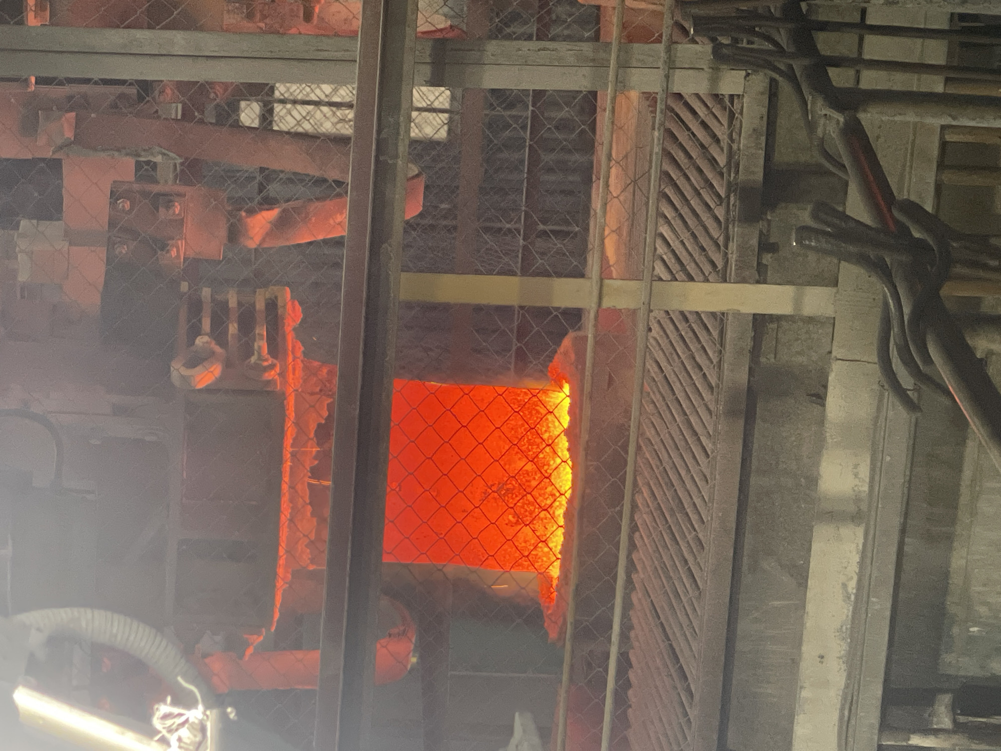 The image depicts an industrial setting, likely a furnace or foundry, with a large volume of intensely glowing orange material visible in the center. This material appears molten and is contained within a recessed area, possibly a crucible or mold. The scene is characterized by high temperatures, indicated by the incandescent glow and possible heat haze or steam.
Surrounding the central glowing material are various industrial structures and equipment. Metal grates and platforms are present, suggesting a multi-level industrial facility. There are also metal railings and mesh fencing, which could serve as safety barriers. Various pipes and conduits are visible, consistent with an industrial process. The overall environment appears somewhat grimy and industrial, with dust or soot likely present on surfaces.
The image does not contain any discernible people or clear indications of human activity within the frame itself, though the presence of tools or implements in the foreground suggests they are used in this environment. The lighting is dominated by the intense orange glow of the molten material, with some ambient light from artificial sources, possibly overhead lamps.
The location provided is the city of Oskemen, Kazakhstan. This context suggests the image was taken at an industrial facility within this city, potentially related to metallurgy, manufacturing, or energy production, given the nature of the scene.