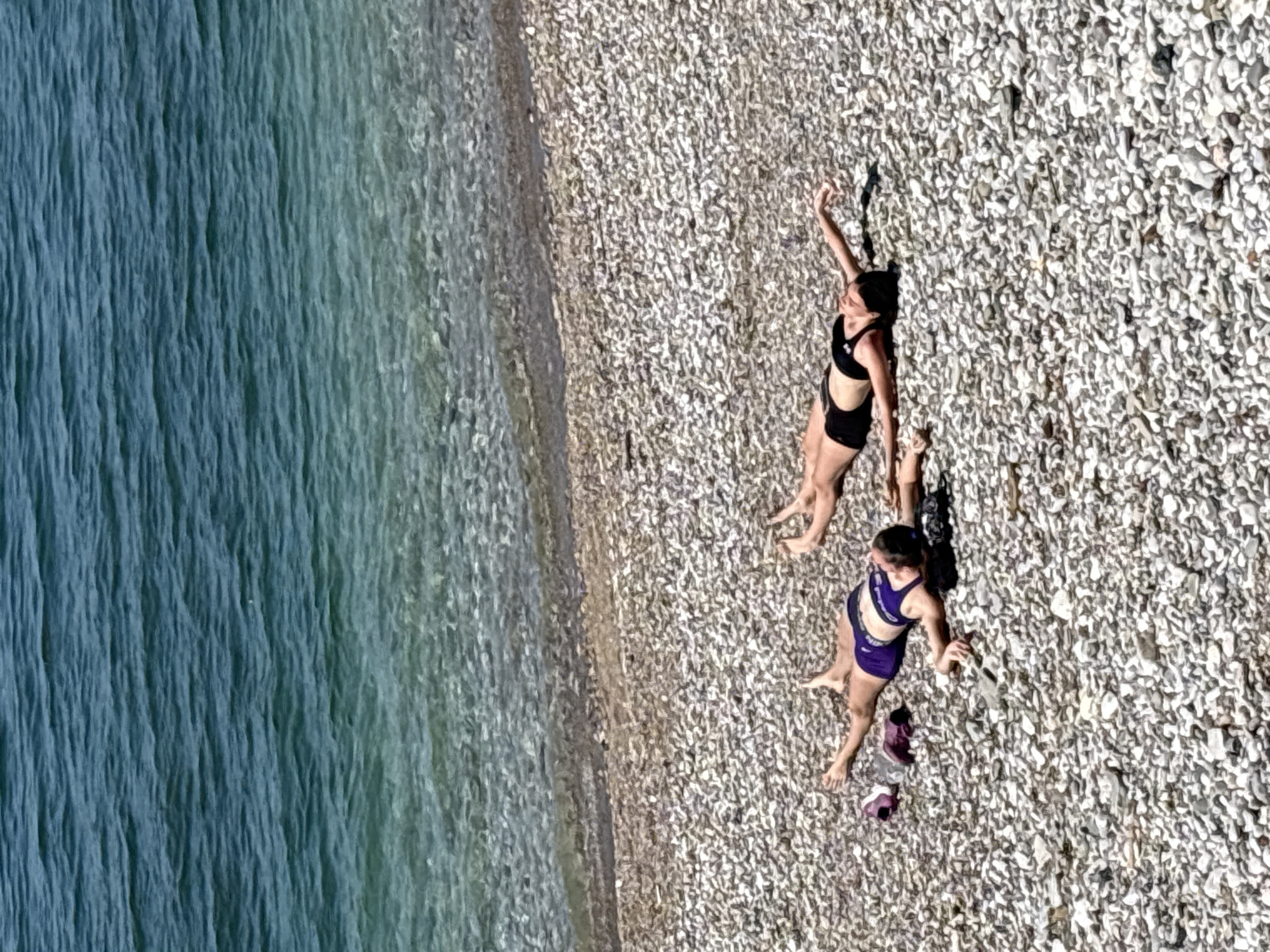 This aerial or high-angle photograph captures a pebble beach in Bar, Montenegro, beside clear blue-green water under bright daylight. The scene features two women relaxing on the light-colored pebbly shore. Strong shadows suggest sunny weather, likely midday or afternoon.
One woman, wearing a black athletic two-piece, is seen lying or reclining on her back with one arm raised. The second woman, dressed in a purple athletic top and matching shorts, is seated or resting on the pebbles nearby, with a pair of purple shoes visible beside her. The top worn by the woman in purple displays a "Nike" logo.
The water, appearing textured from currents or small waves, transitions to the beach through a darker band of potentially wet pebbles. The overall atmosphere is one of serene leisure, with the individuals enjoying the coastal environment. The distinct white and gray pebbles of the beach contrast with the vibrant blues and turquoises of the sea, offering a unique perspective of this coastal relaxation by the Adriatic Sea.
