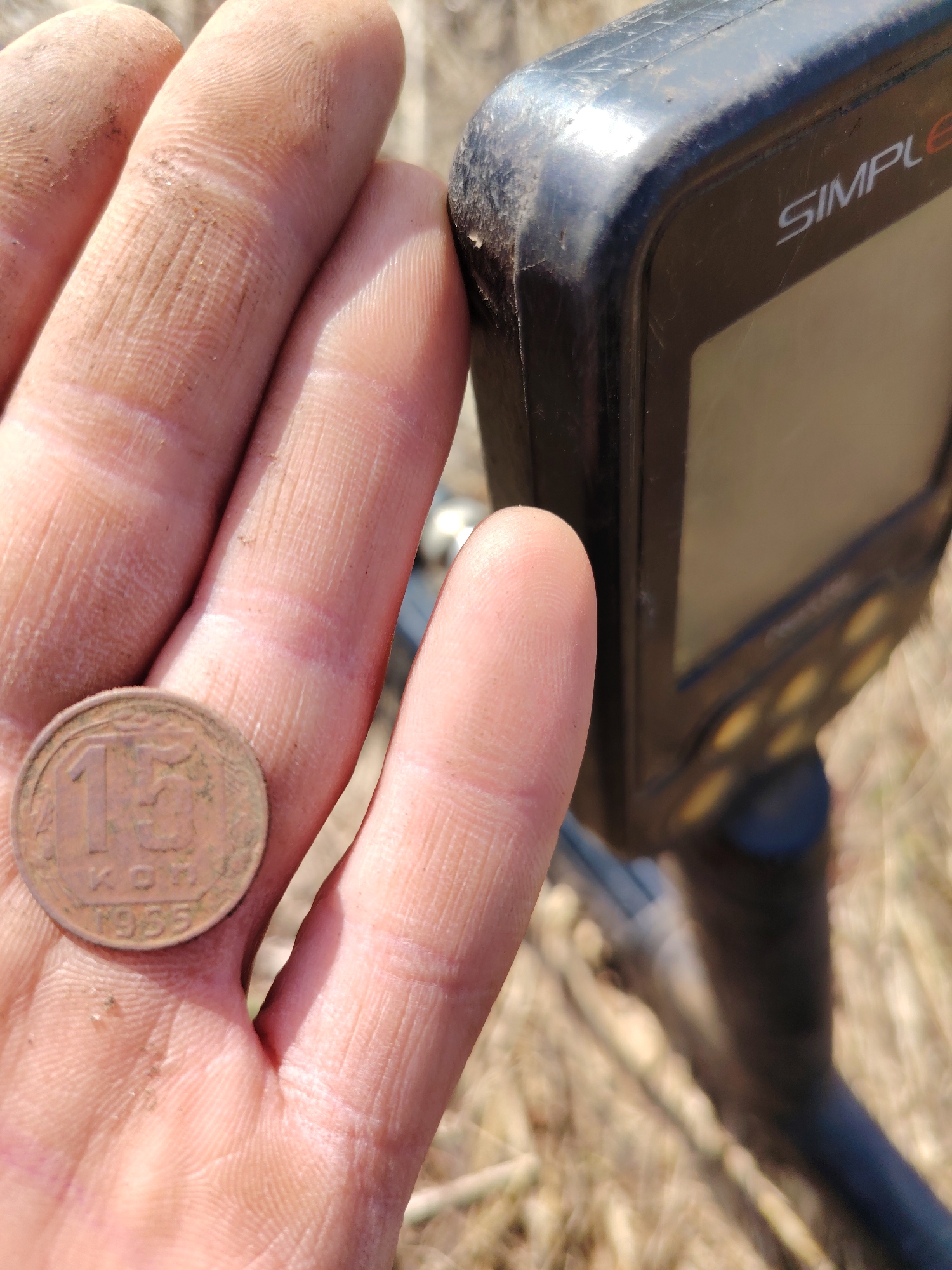 A close-up shot captures a dirt-stained hand holding an old coin, with a metal detector partially visible in the background. The hand is covered in light brown soil and dust, particularly on the fingertips and palm, suggesting recent interaction with earth.
The coin, dull reddish-brown in color, is positioned in the center of the palm. Clearly visible on its surface are the large numbers "15", followed by the Cyrillic abbreviation "КОП." (indicating Kopeks), and the year "1955". This identifies it as a Soviet-era 15 Kopek coin from 1955.
To the right of the hand, the black control unit of a metal detector is in sharp focus. The brand name "SIMPLE" is partially visible on its casing, likely indicating a "Nokta Makro Simplex" model. The detector's surface shows signs of wear, with scratches and dirt, consistent with outdoor use. A dark screen and some yellow or gold-colored buttons are visible. The overall setting appears to be outdoors, in a field or grassy area, with blurred, dry, light-colored vegetation in the background. The bright lighting suggests it is daytime. The image depicts a successful find during a metal detecting activity.